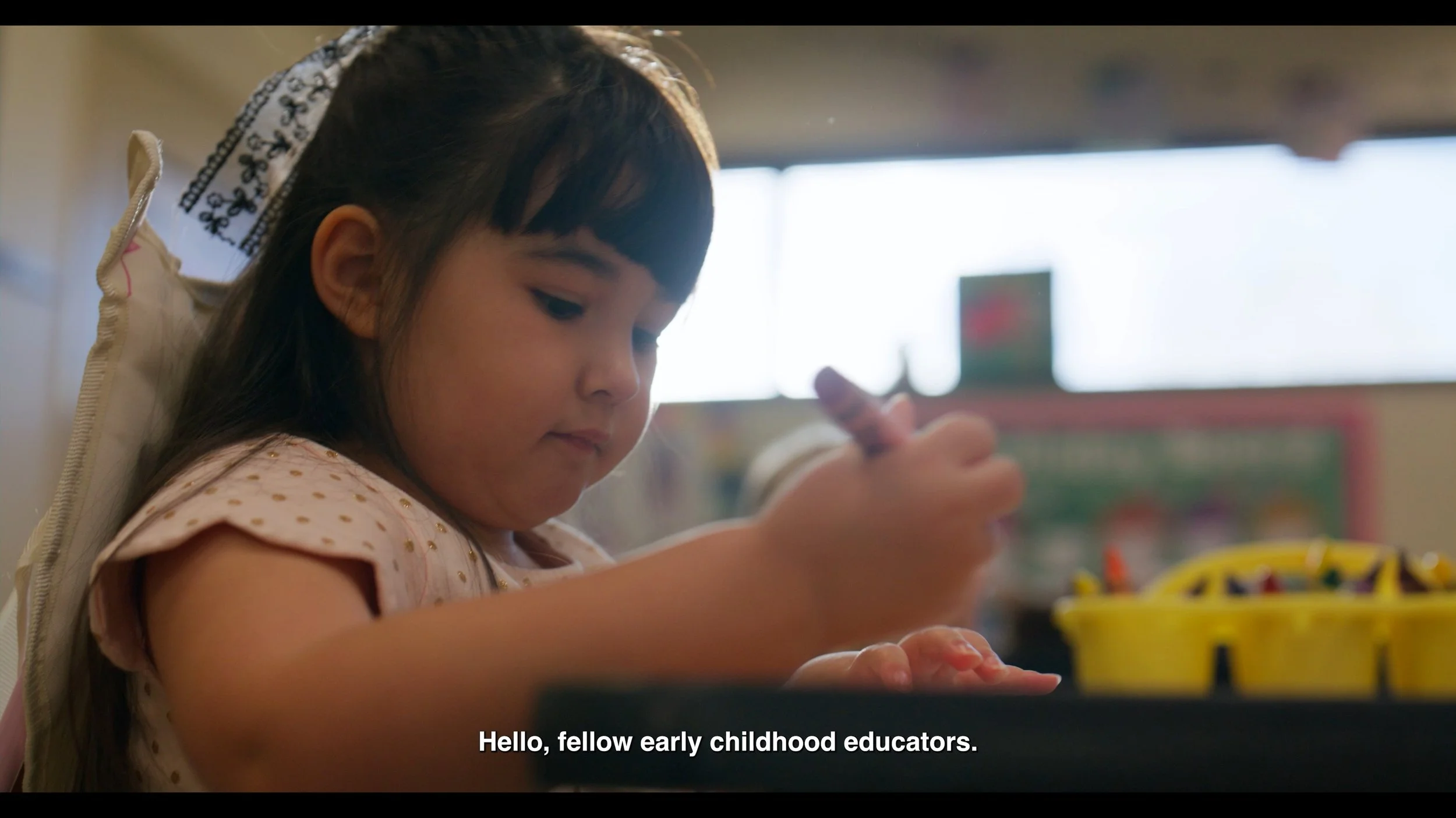 A young girl with dark hair and a white headband, sitting at a table with a yellow container of markers, holding a marker in her right hand and drawing. She is wearing a light-colored shirt with gold polka dots. The background shows a bright classroo