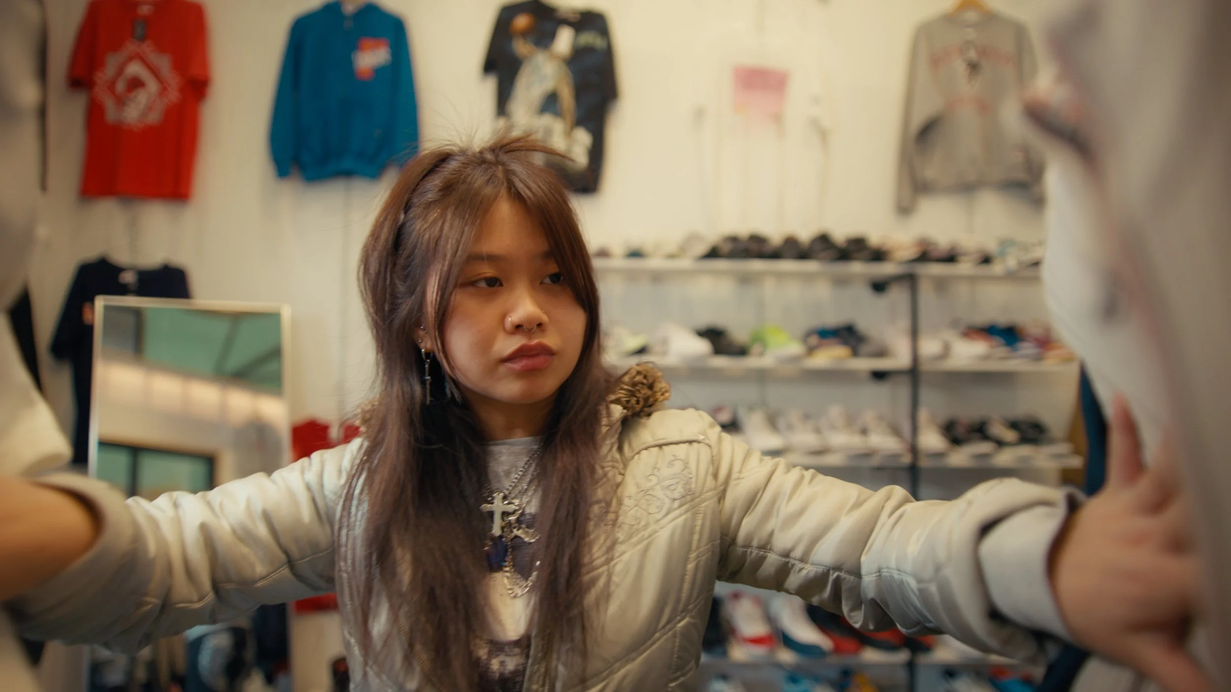 A young woman with long brown hair and earrings is shopping in a sneaker store, holding a shoe in each hand and examining them.