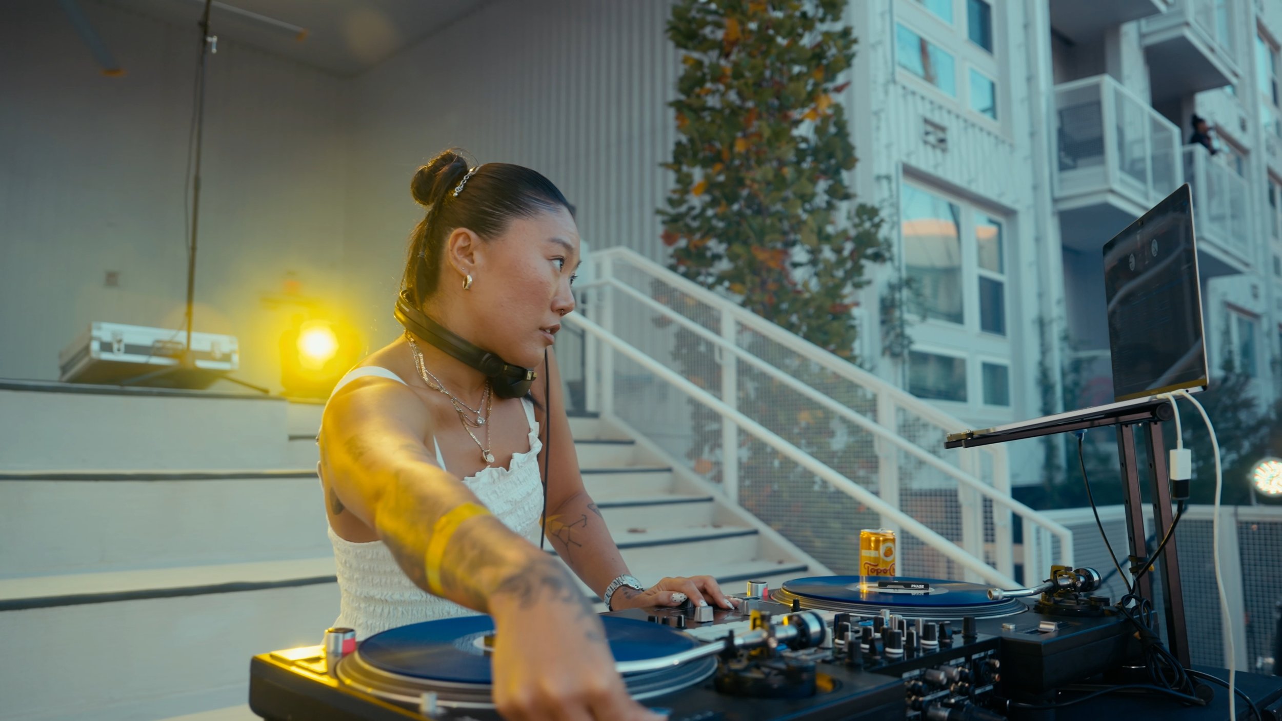A female DJ with tattoos and jewelry operating a turntable setup outdoors during sunset, with a laptop and a can of Tito's vodka nearby.