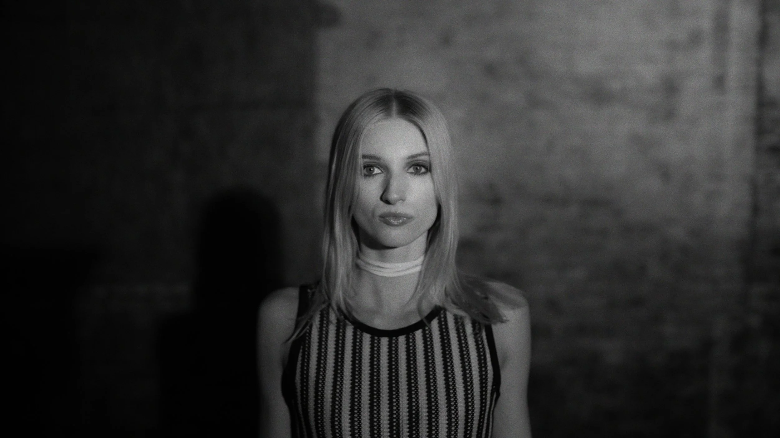 A black and white portrait of a young woman with shoulder-length hair, wearing a sleeveless striped top and a choker, standing against a textured wall.