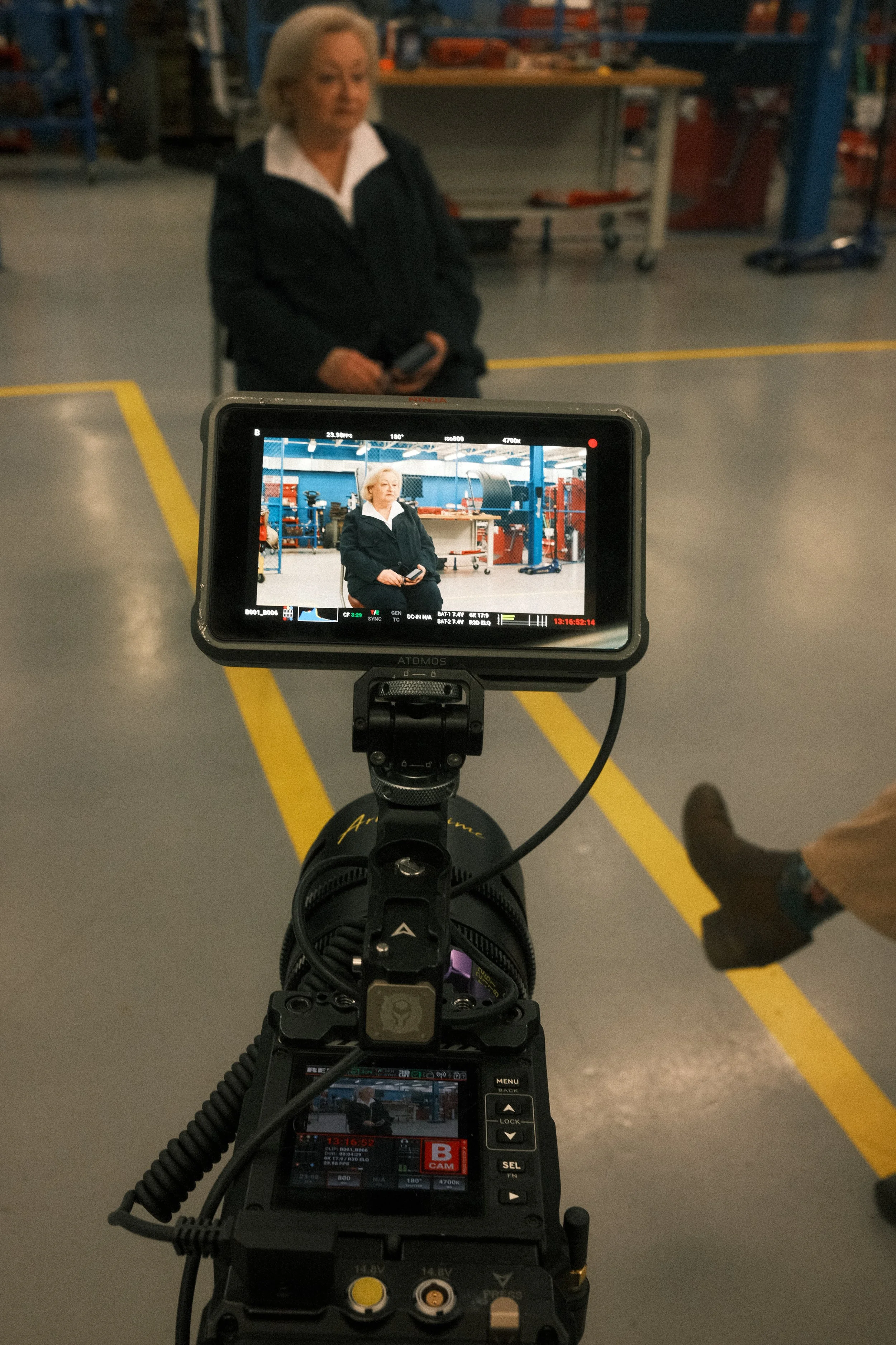 A woman with short blonde hair wearing a black jacket and white blouse sitting in a large industrial warehouse, seen through the screen of a professional camera mounted on a stabilizer.