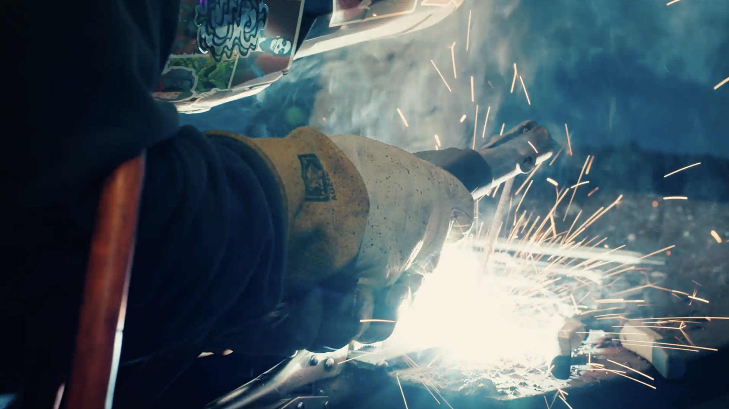 A worker wearing gloves and a welding helmet is welding metal, creating sparks and bright light.