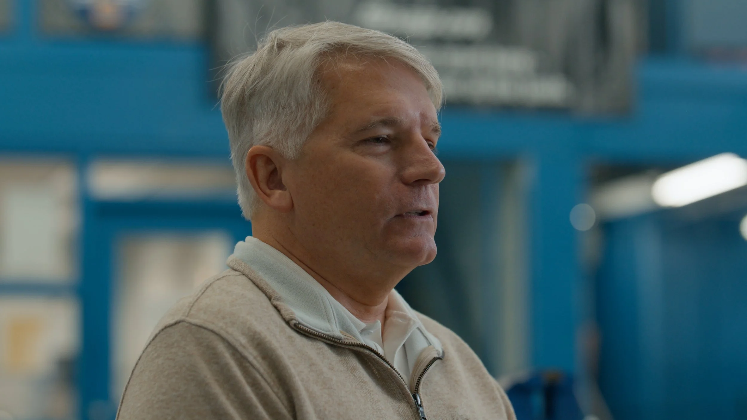 A middle-aged man with gray hair wearing a beige zip-up sweater in an indoor setting with blue structural elements in the background.