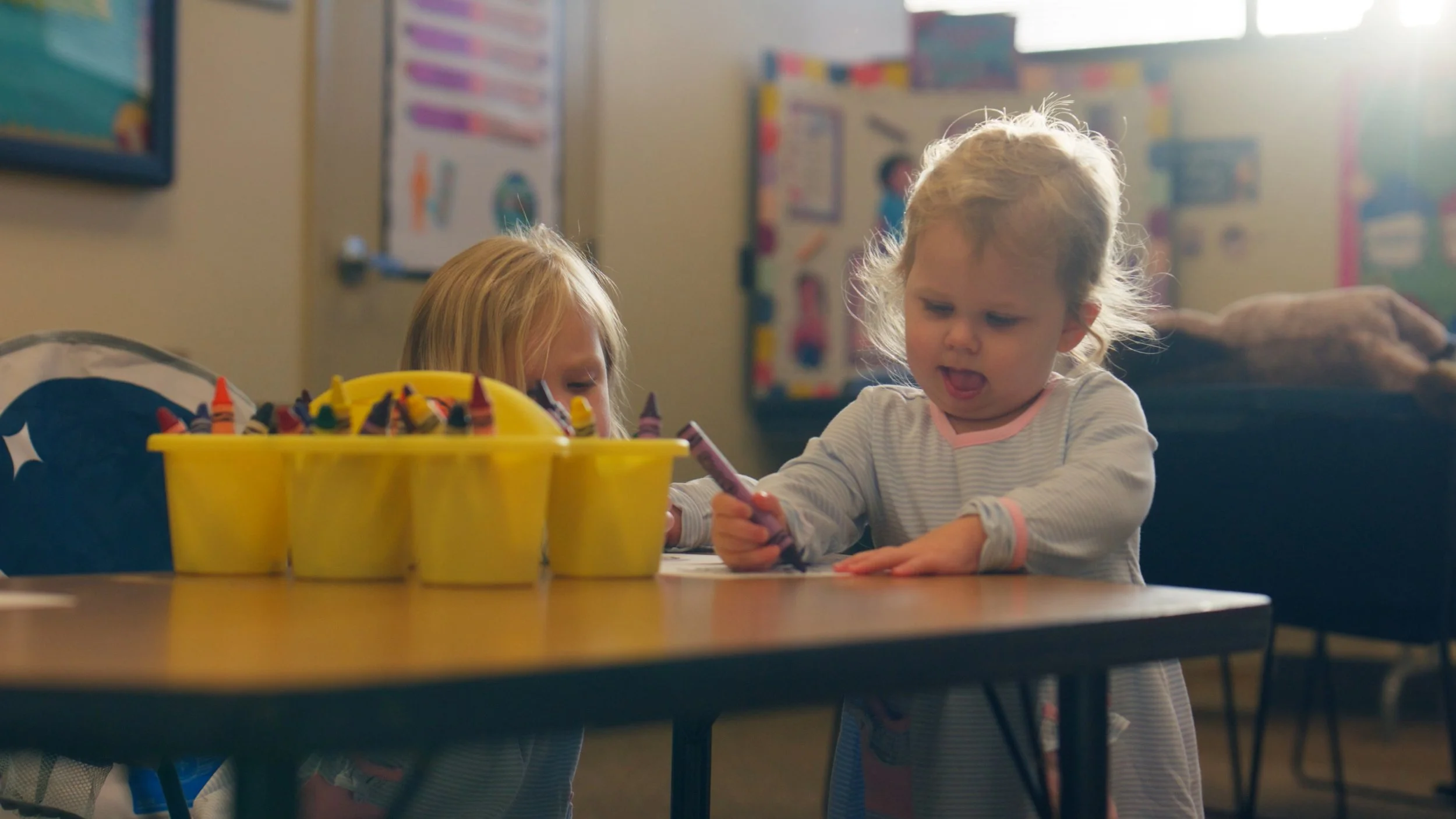 Two young girls with blonde hair sitting at a table in a classroom, coloring with crayons. One girl appears confused or upset. A yellow container filled with crayons is on the table.
