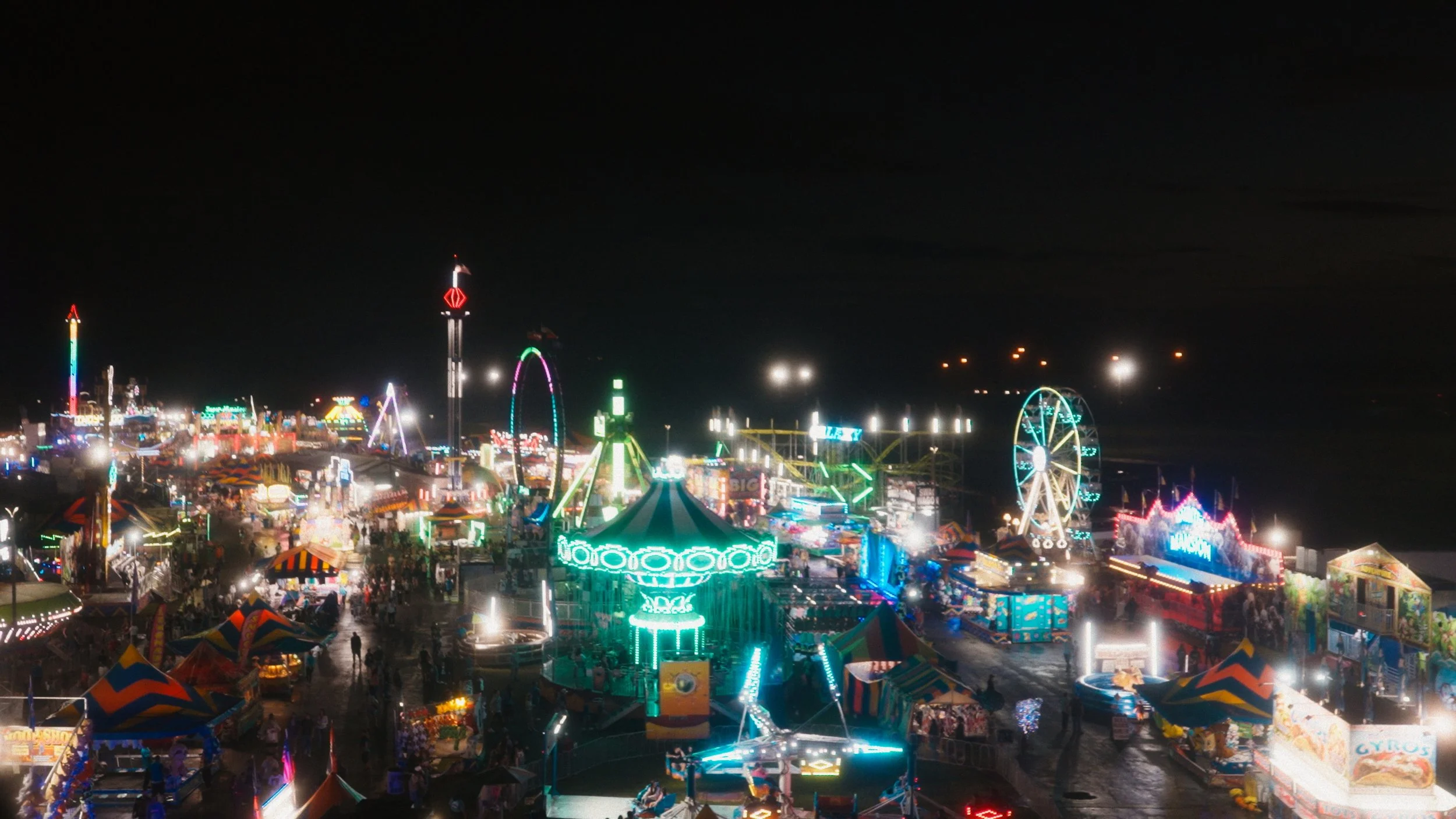 Night scene of a colorful carnival with rides, tents, and bright lights.