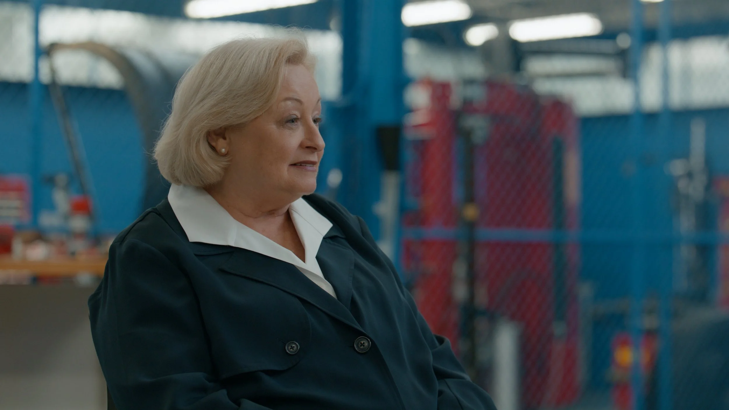 An elderly woman with blonde hair, wearing a navy blazer and white shirt, standing in what appears to be a warehouse or industrial setting with blue fencing and red equipment in the background.