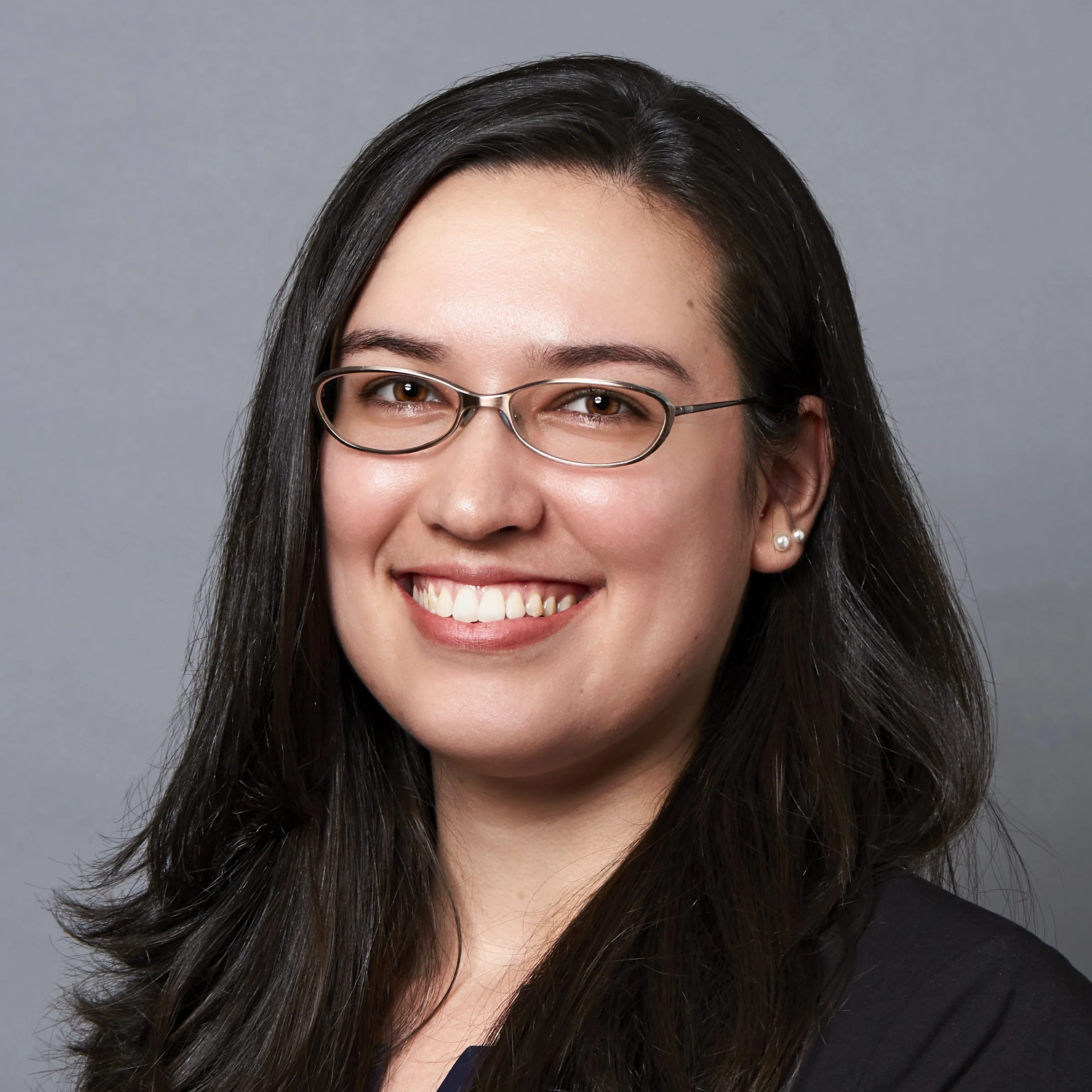 Three-quarter portrait of Melanie Iglesias in front of a gray background. A white woman with dark long hair, brown eyes, brown metal glasses, and pearl earrings.
