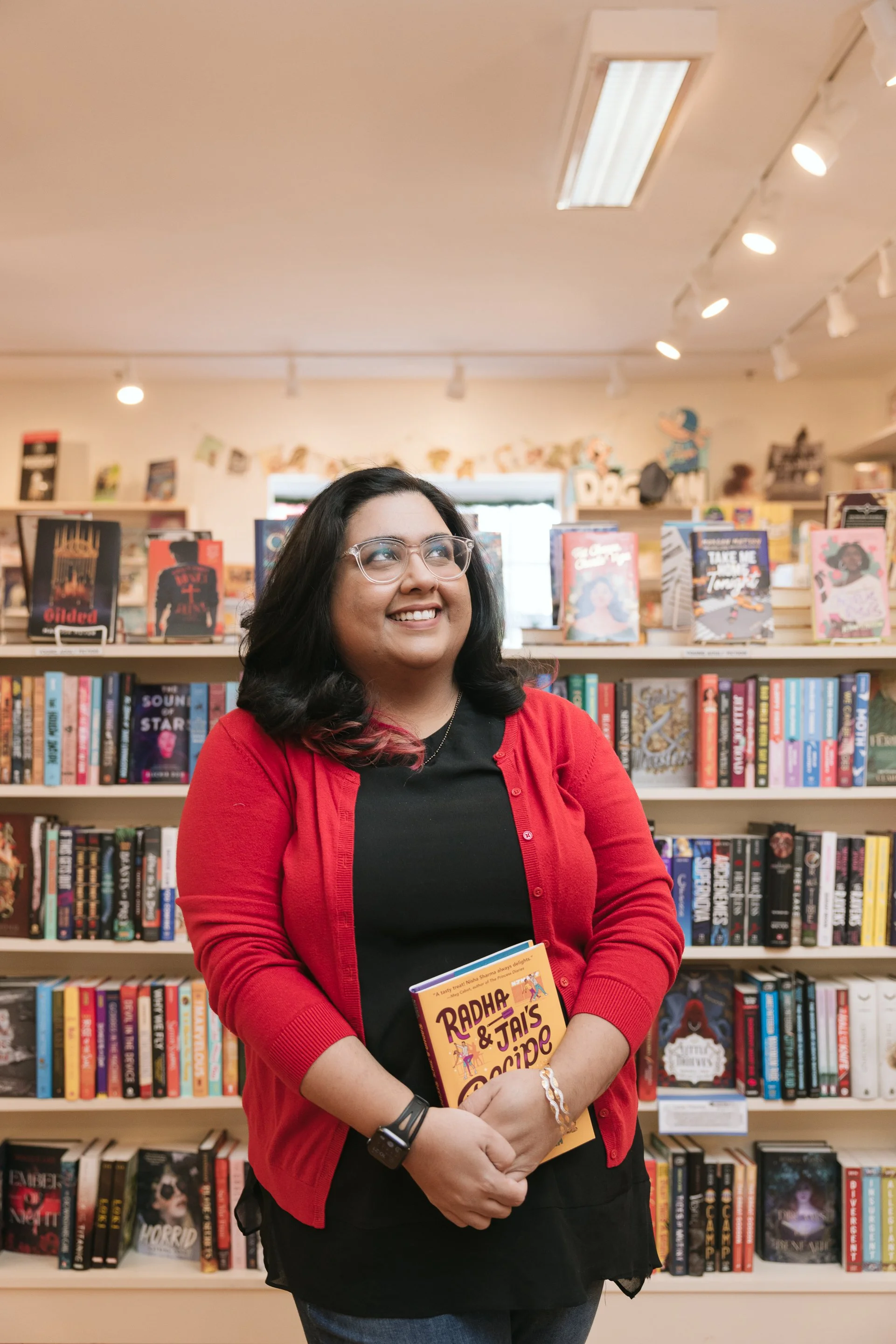 Author Nisha Sharma is standing in a bookstore while wearing a red cardigan over a black blouse and jeans while holding a copy of her Y.A. novel RADHA & JAI'S RECIPE FOR ROMANCE.