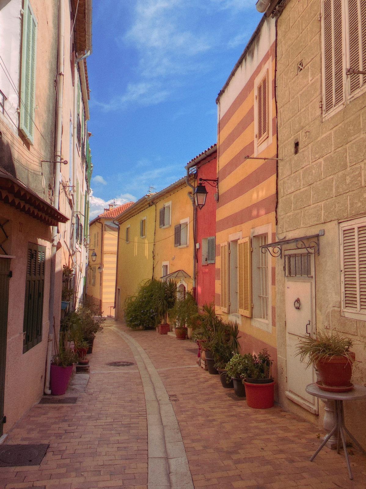 Narrow cobblestone street lined with colorful houses decorated with flower pots, under a blue sky.