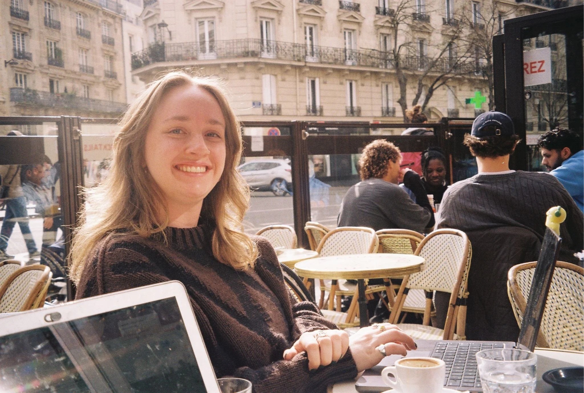 Young woman smiling at a café table with laptops, coffee, and water, with other customers dining outside and city buildings in the background.