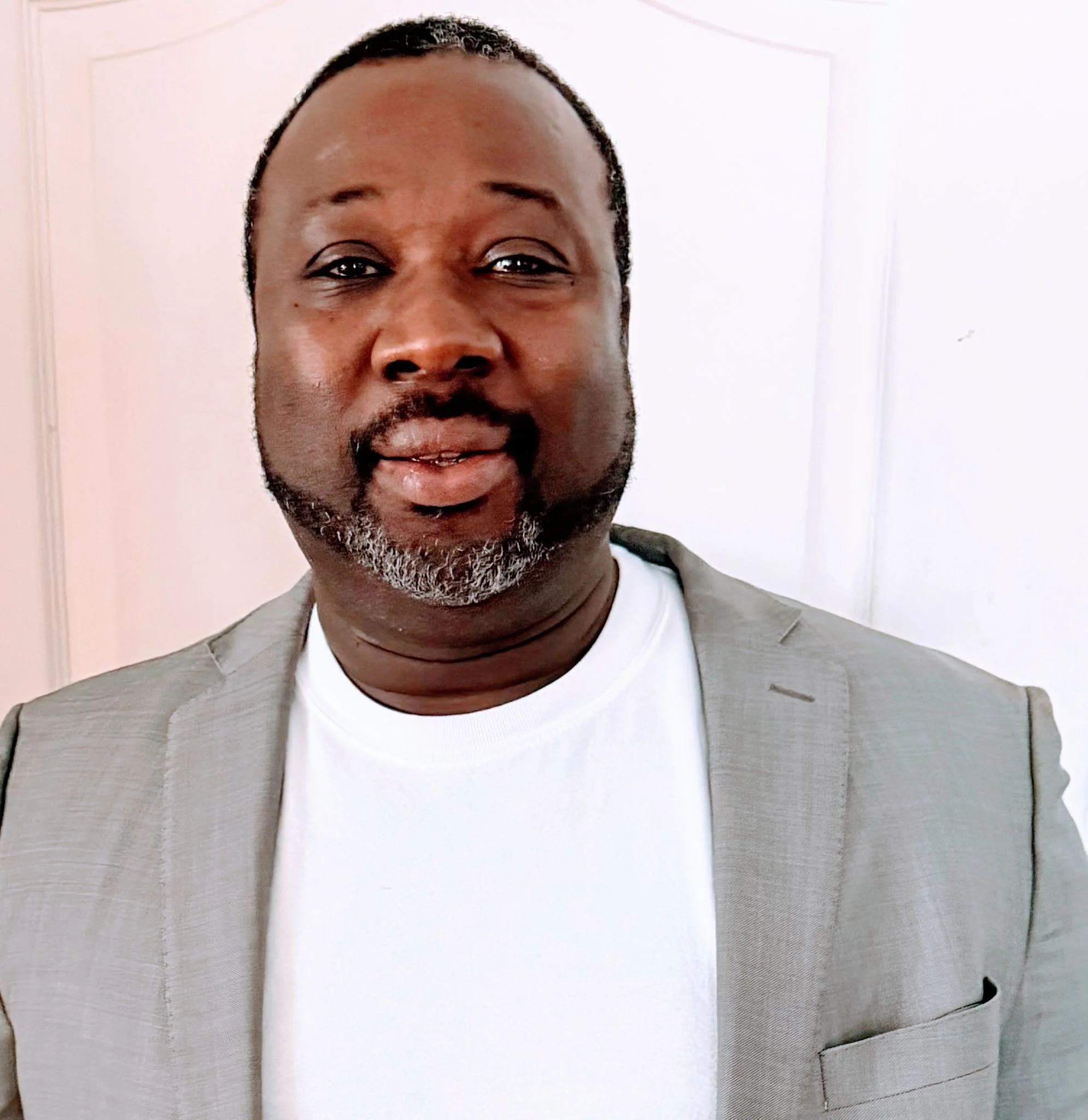 Homme portant un costume gris clair avec t-shirt blanc, souriant devant un fond blanc.