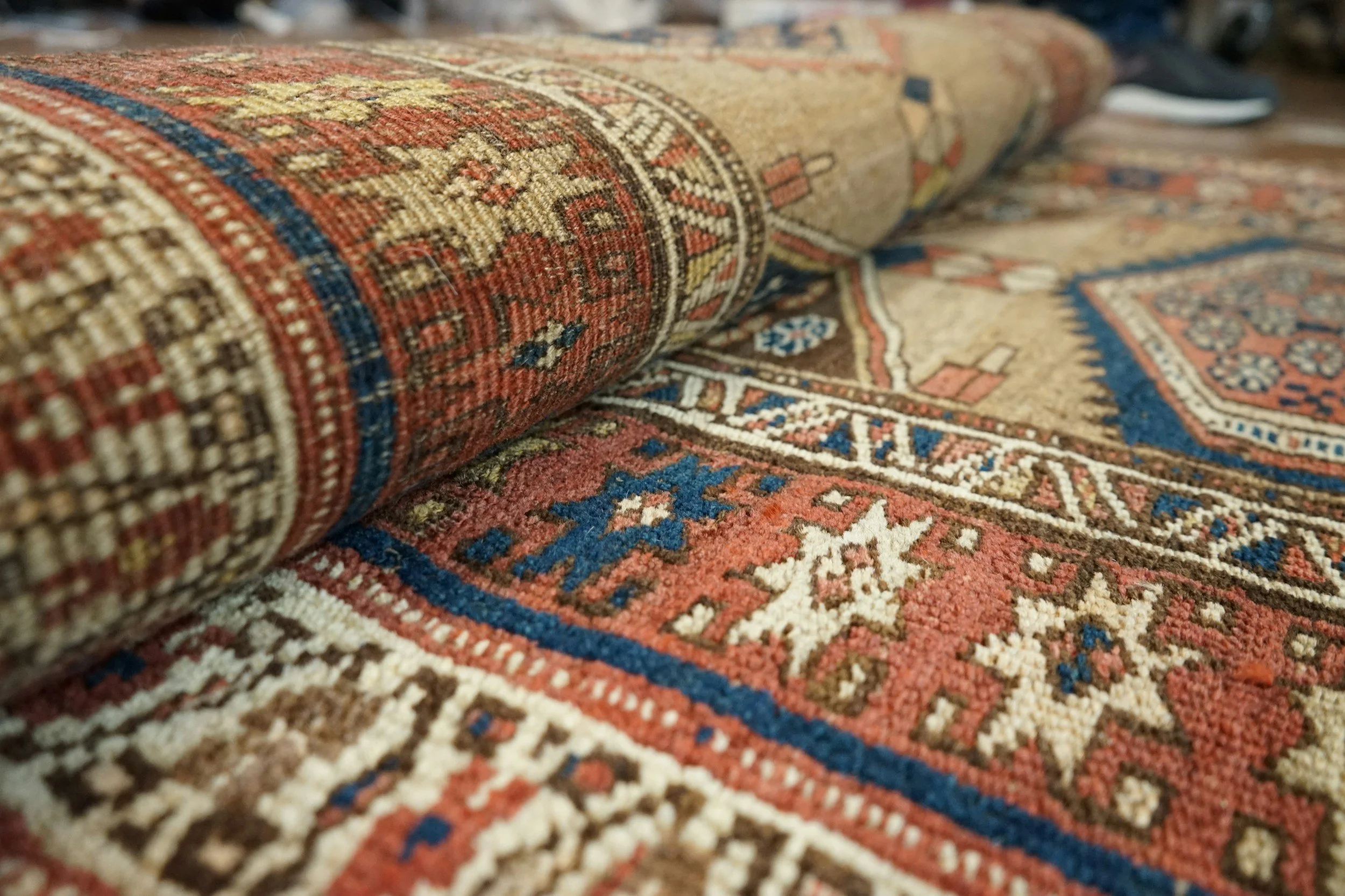 Close-up of a rolled Persian-style carpet with intricate patterns in red, blue, beige, and brown, lying on a wooden floor.