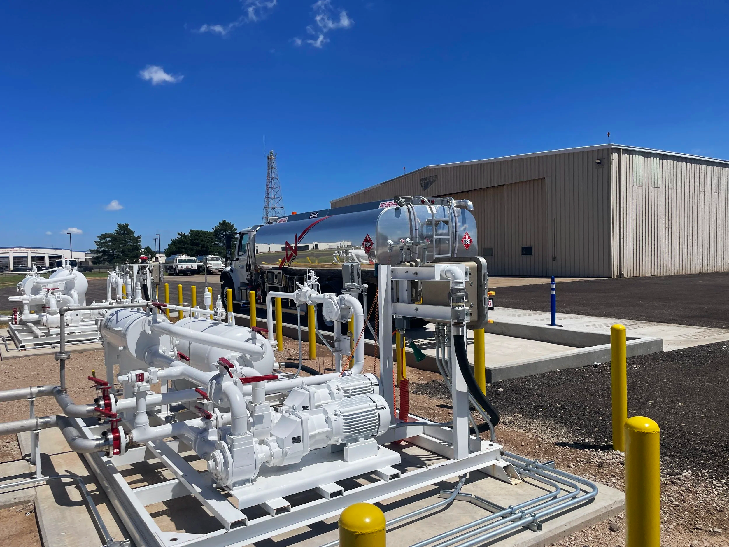 A large industrial fuel or chemical storage tank on a truck, surrounded by pipes and valves, in an outdoor industrial area with a beige building and a communication tower in the background under a clear blue sky.