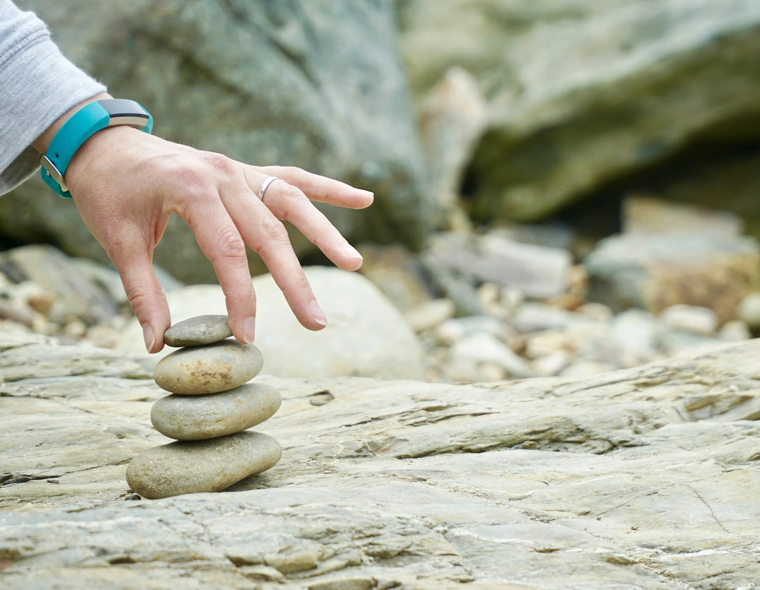 A hand with a wedding ring and a blue wristband, balancing five stones in a vertical stack on a rocky surface in an outdoor setting.