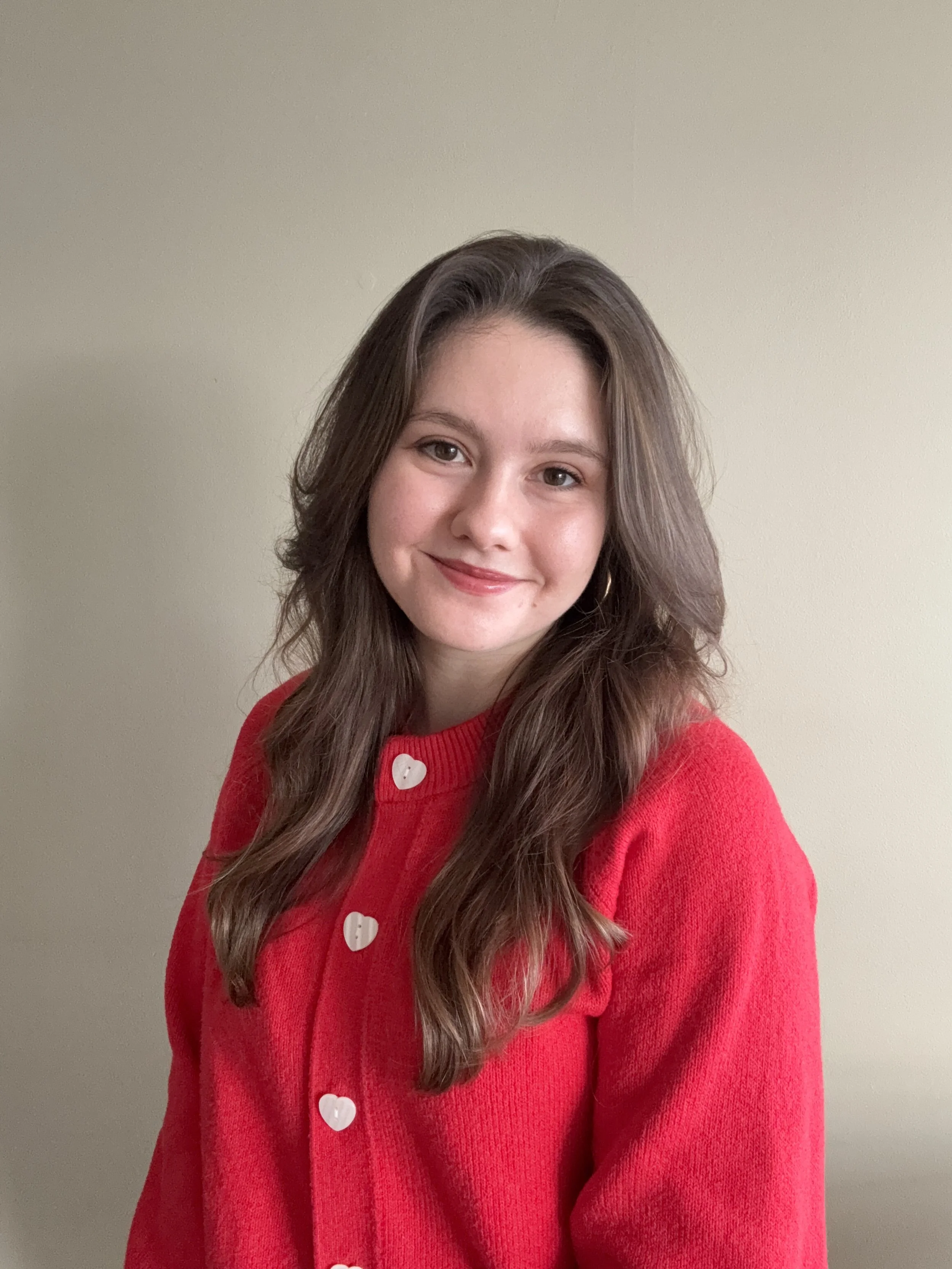 A young woman with long, wavy brown hair and fair skin, smiling, wearing a red sweater with white heart-shaped buttons, standing against a plain beige wall.