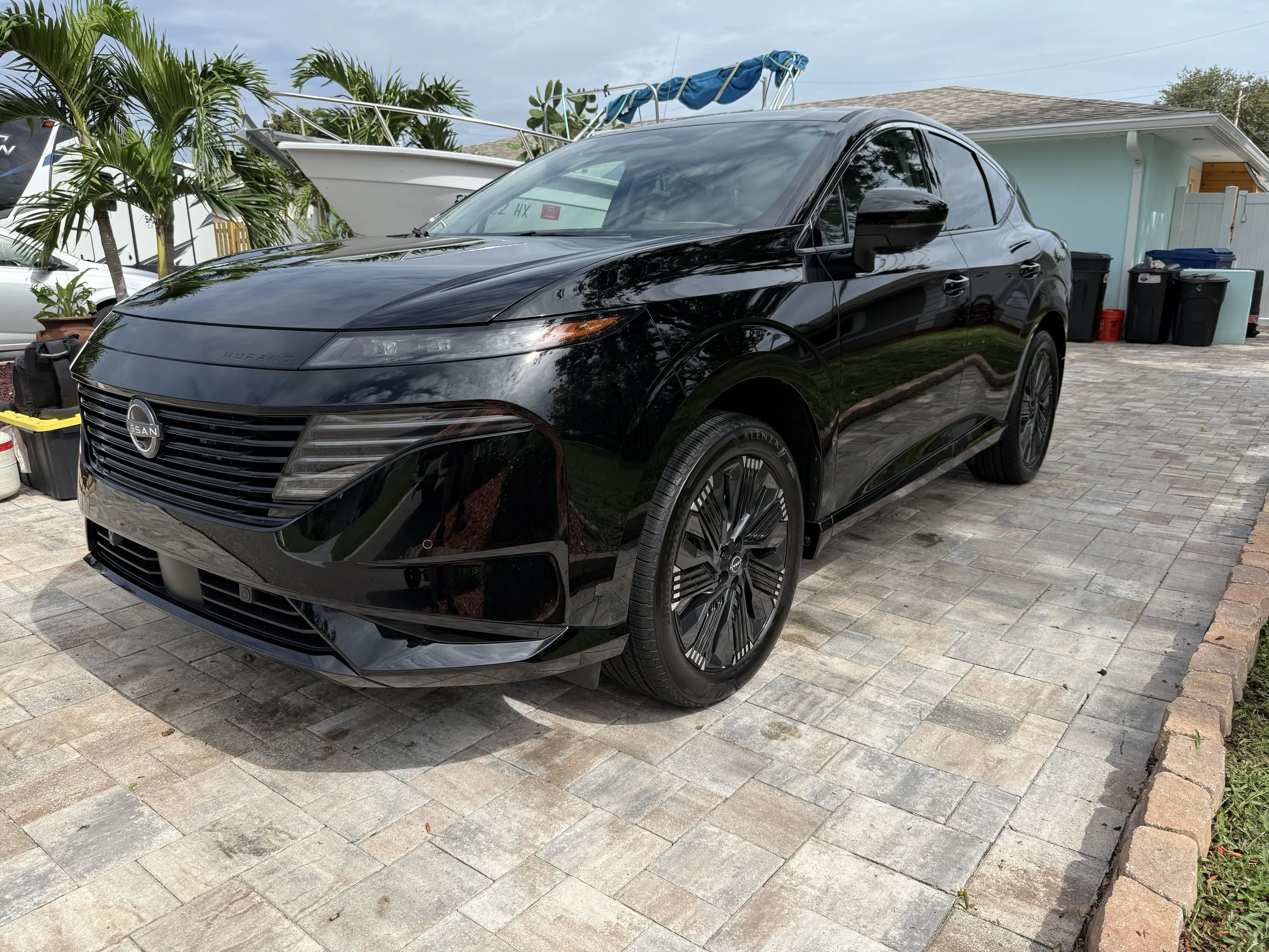 A sleek black electric Nissan Murano parked on a paved driveway near a house with a light blue exterior and trash bins. In the background, there are palm trees and a boat on a trailer.