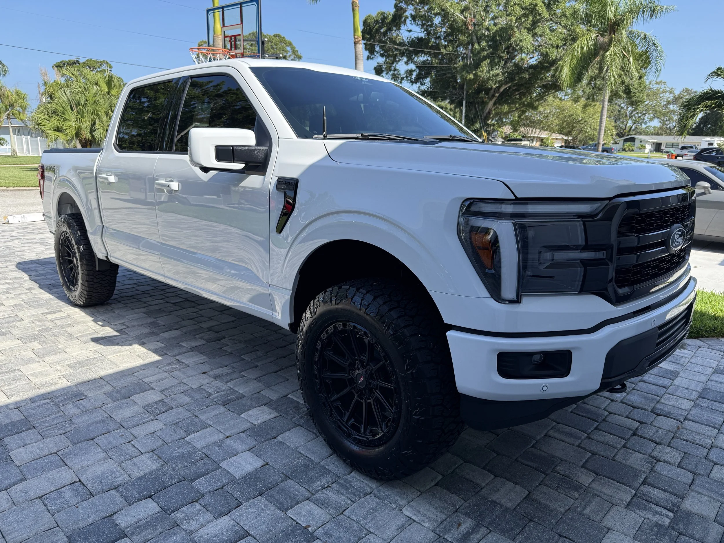 White Ford pickup truck parked on stone pavement with palm trees and a basketball hoop in the background.