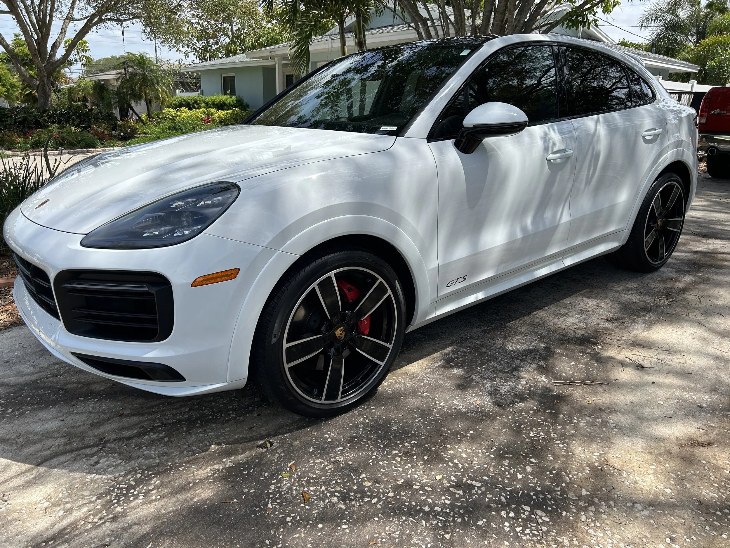 White Porsche Macan GTS parked on a shaded driveway with trees and houses in the background.