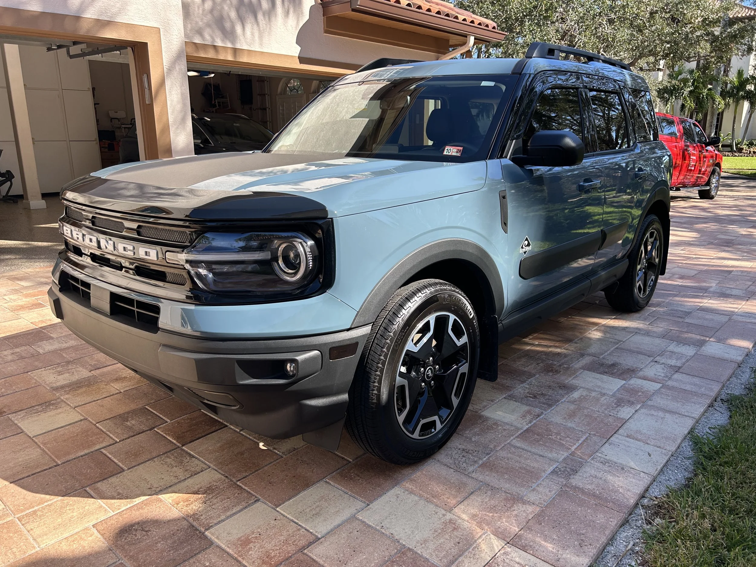 A blue Ford Bronco parked on a brick driveway in front of a house, with a red pickup truck behind it and trees in the background.