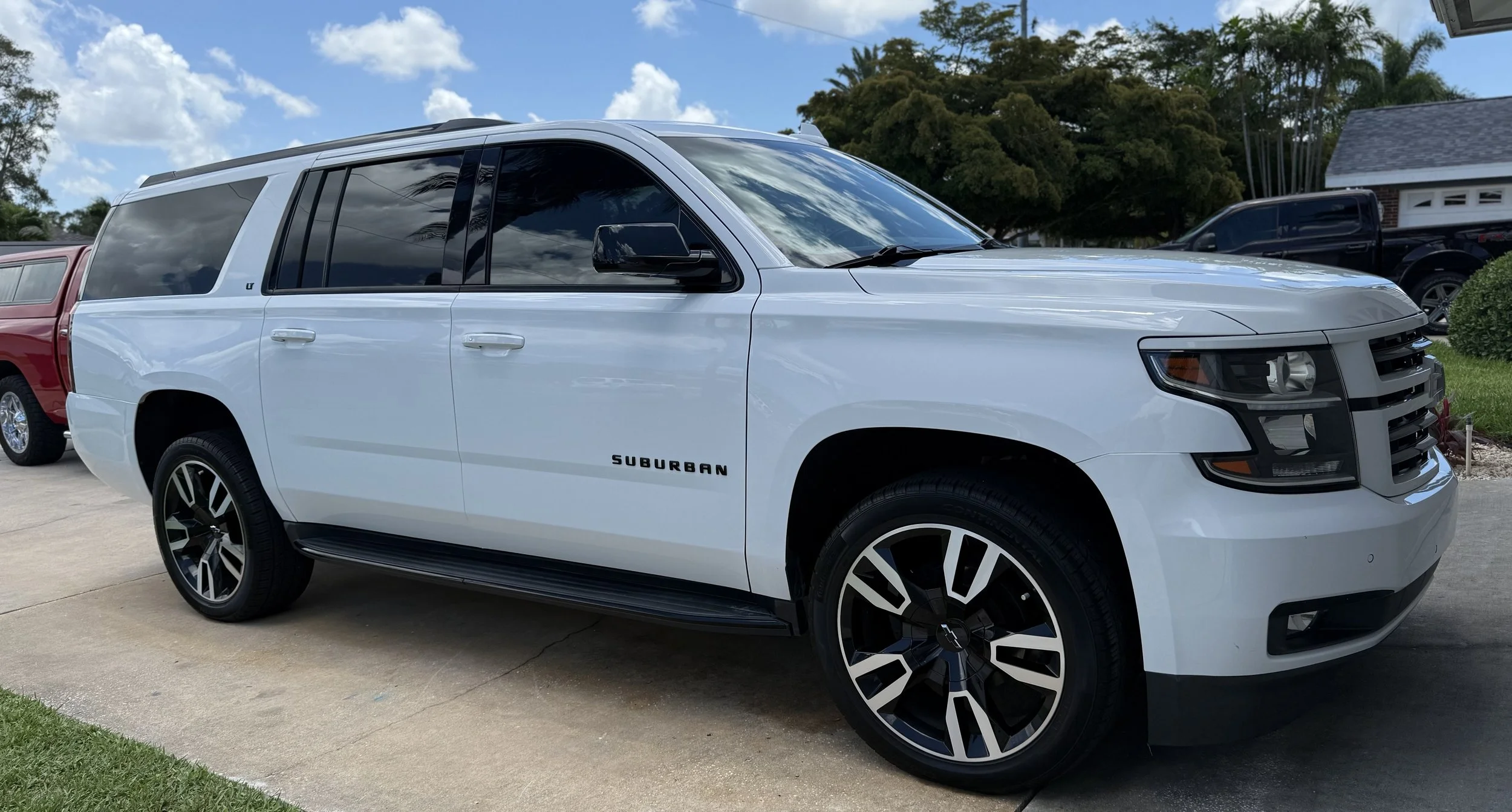 White Chevrolet Suburban SUV parked on concrete driveway with other vehicles and trees in the background.