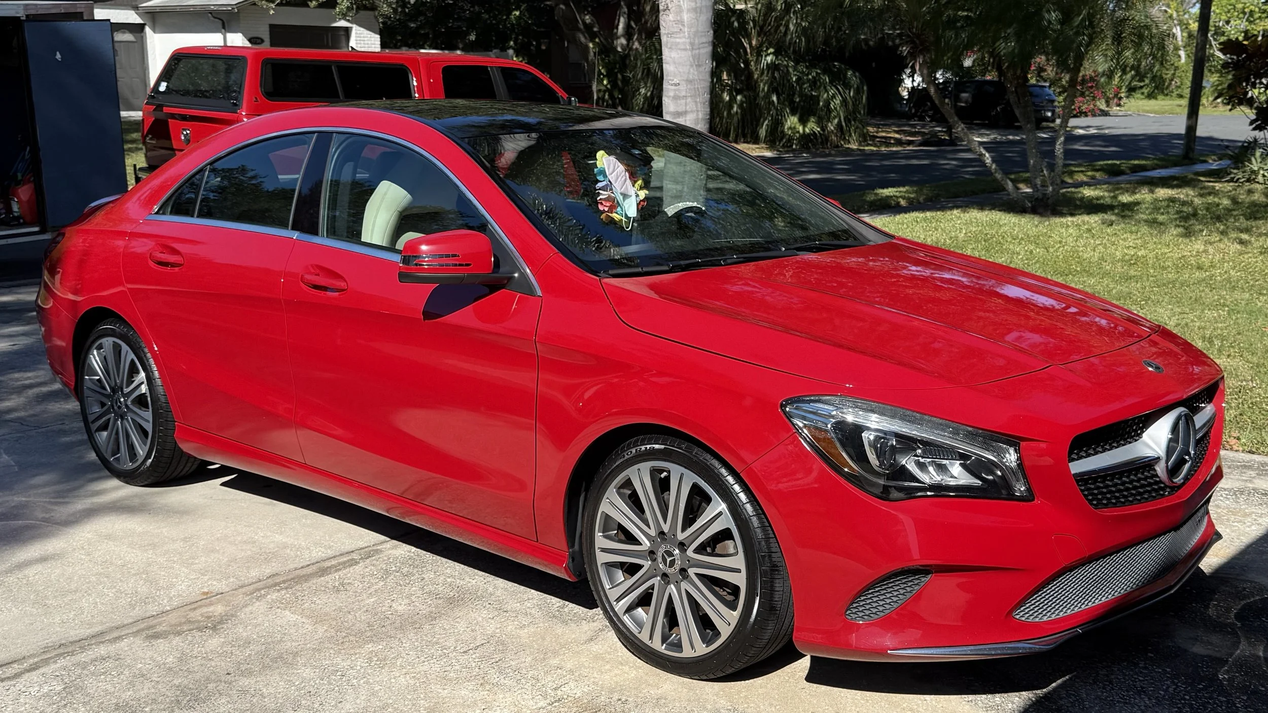 Red Mercedes-Benz sedan parked on driveway with sun reflecting off its surface, with a red SUV and trees in the background.