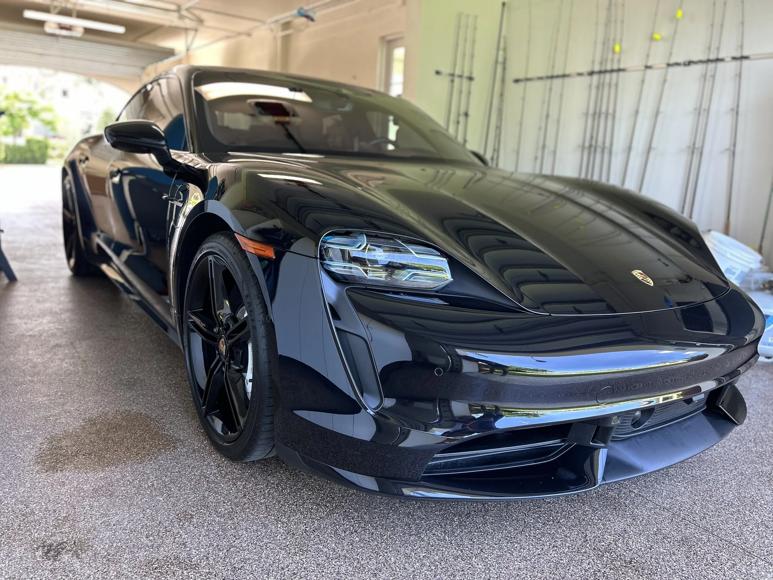 Black Porsche sports car parked inside a garage with a beige wall and green exterior visible through an open door.