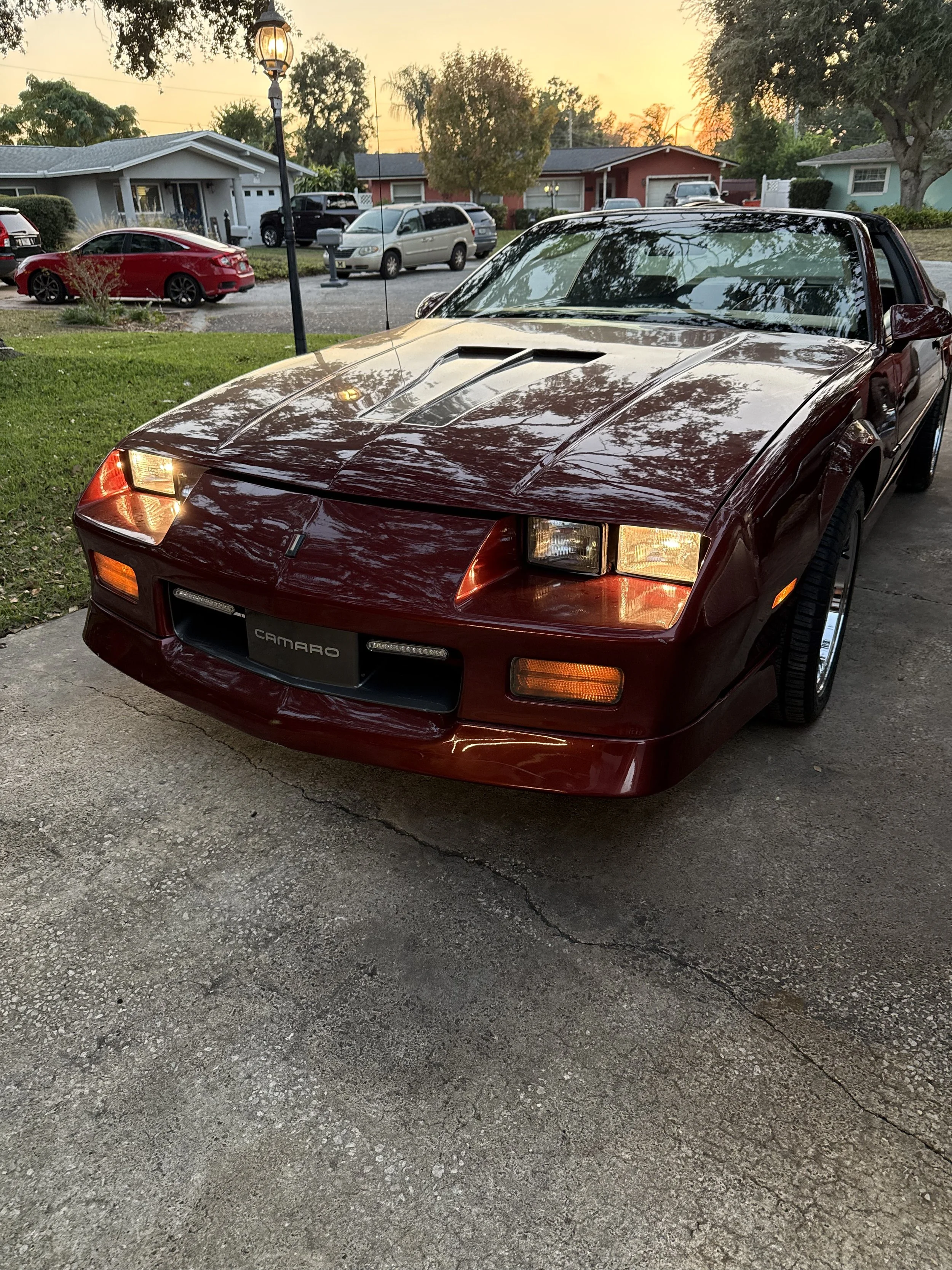 A red Chevrolet Camaro sports car parked on a residential driveway at sunset, with houses, cars, trees, and a lamppost in the background.