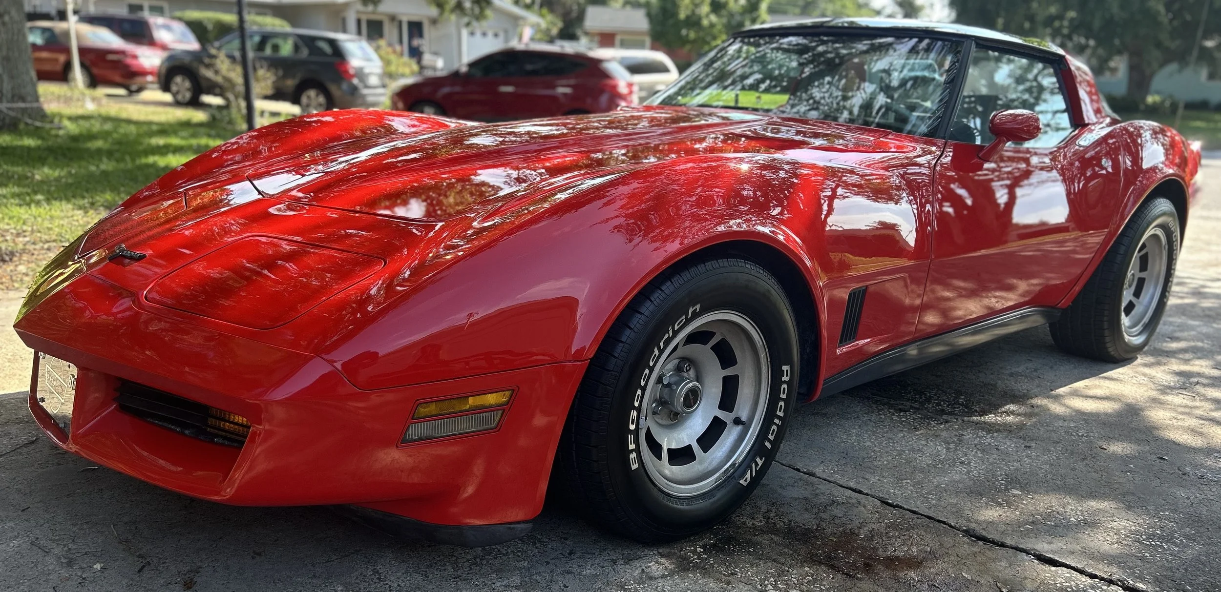 A red vintage Chevrolet Corvette sports car parked on the side of a street, reflecting trees and sky on its shiny surface.