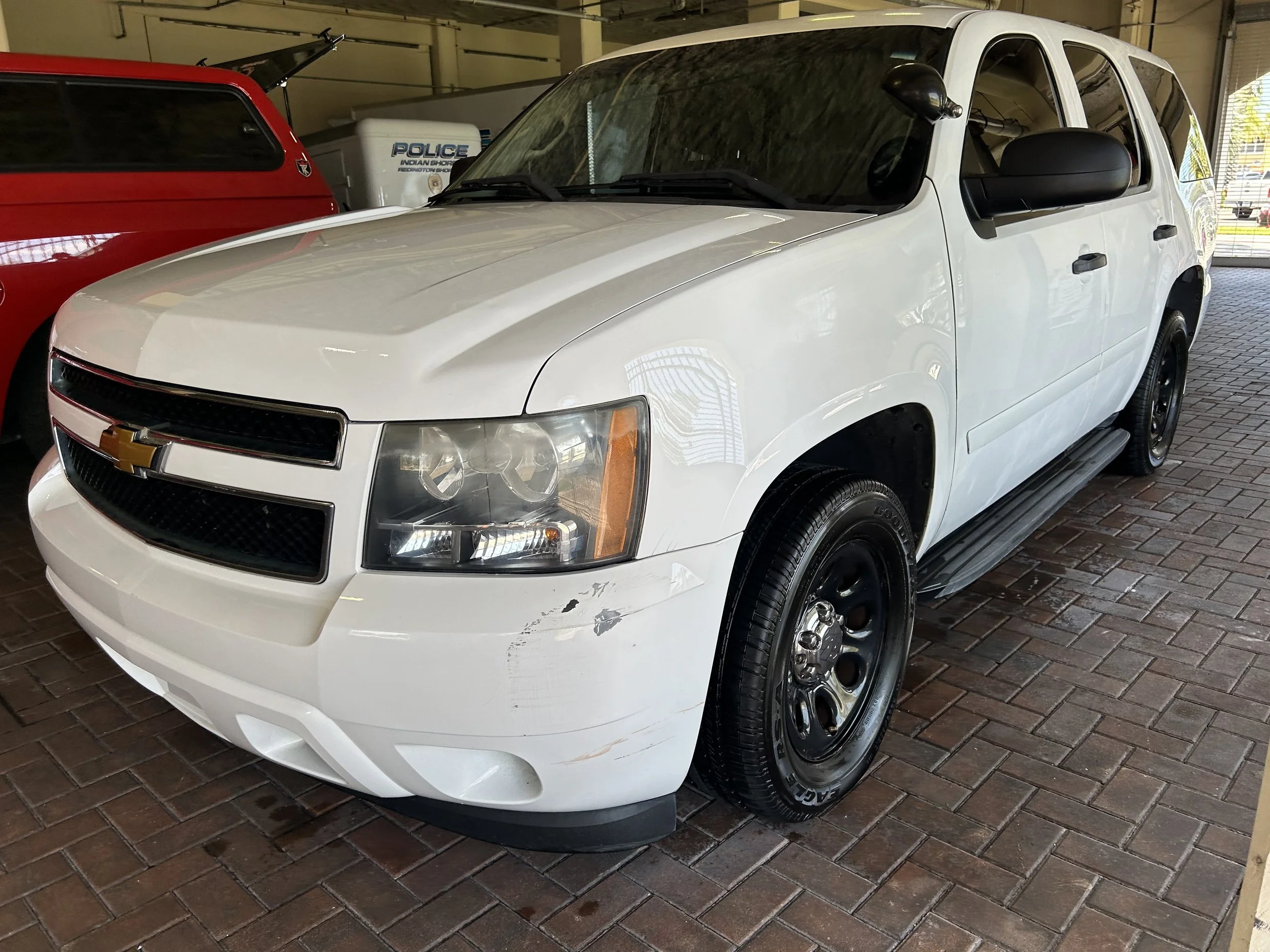 White Chevrolet police vehicle with black wheels parked inside a garage, with damage on the front bumper and scratches on the side.