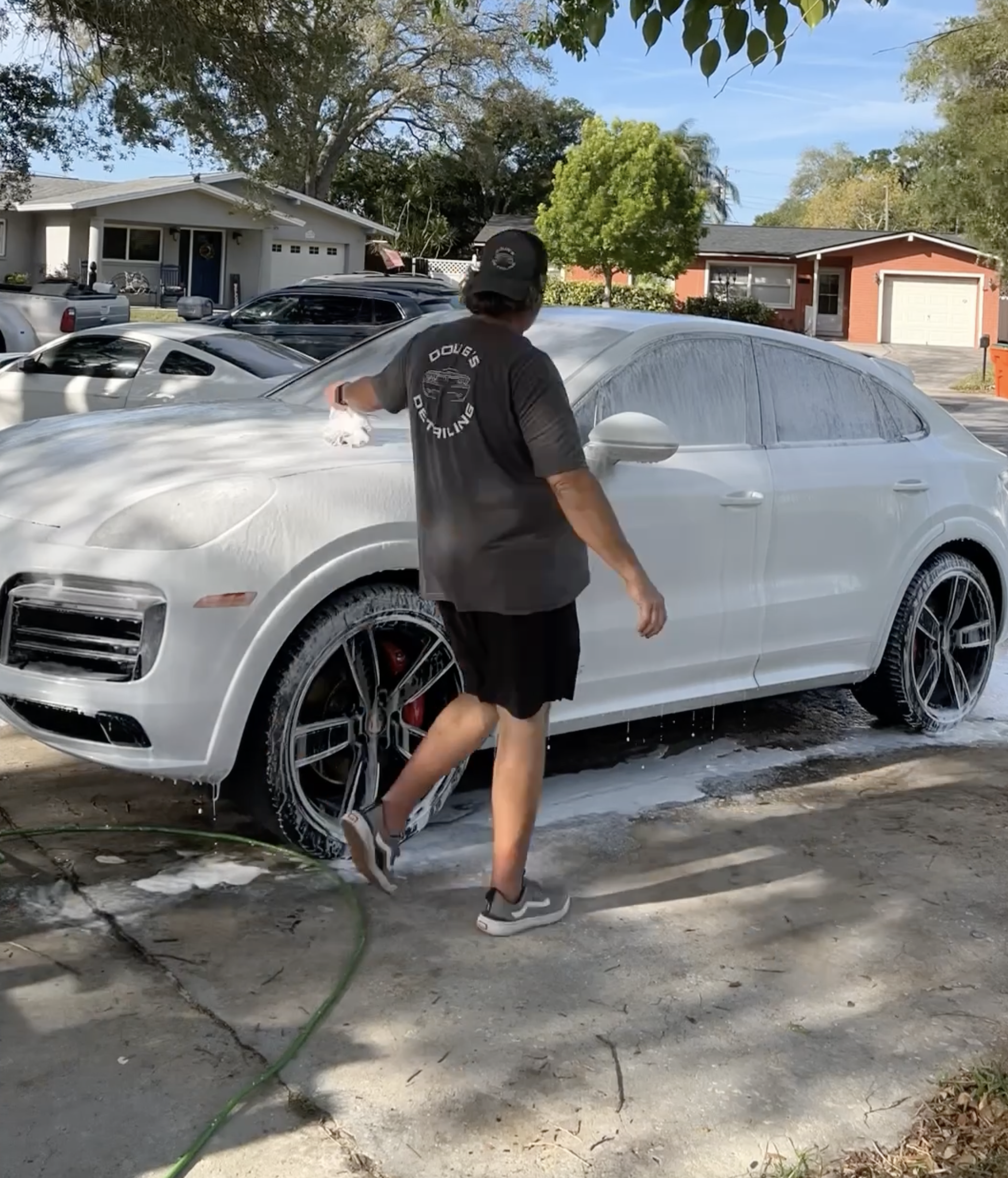 A person is washing a white luxury SUV with foam suds, standing outdoors on a driveway, surrounded by trees and residential houses.