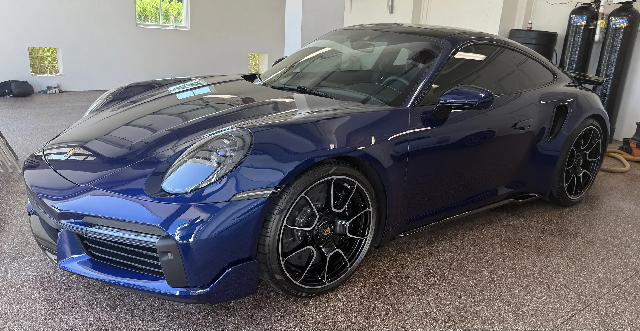 A sleek, blue Porsche sports car parked inside a garage with a textured brown floor, and various tools and storage containers in the background.