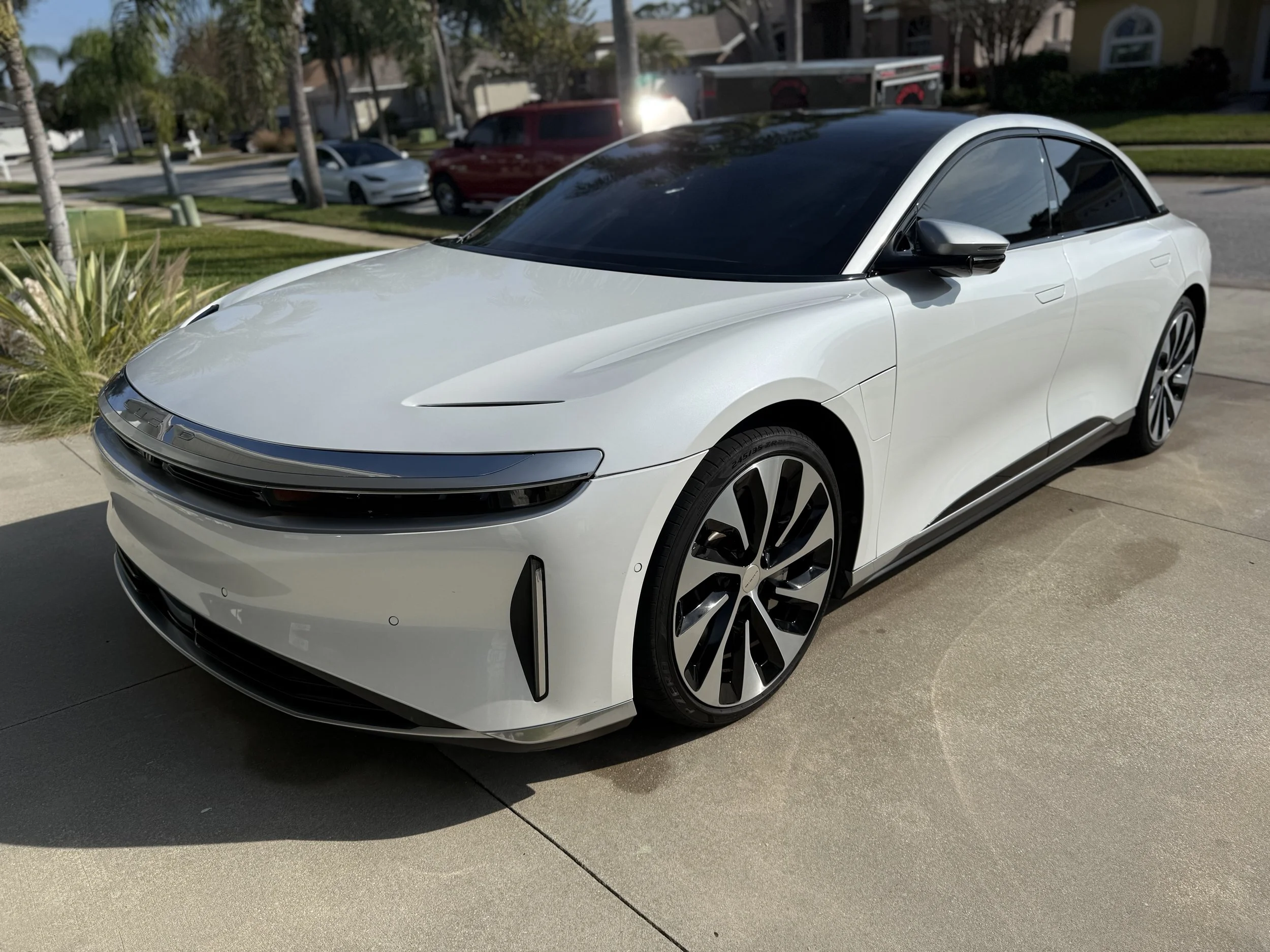 A white electric luxury coupe parked on a driveway in a suburban neighborhood with green lawns, palm trees, and houses in the background.