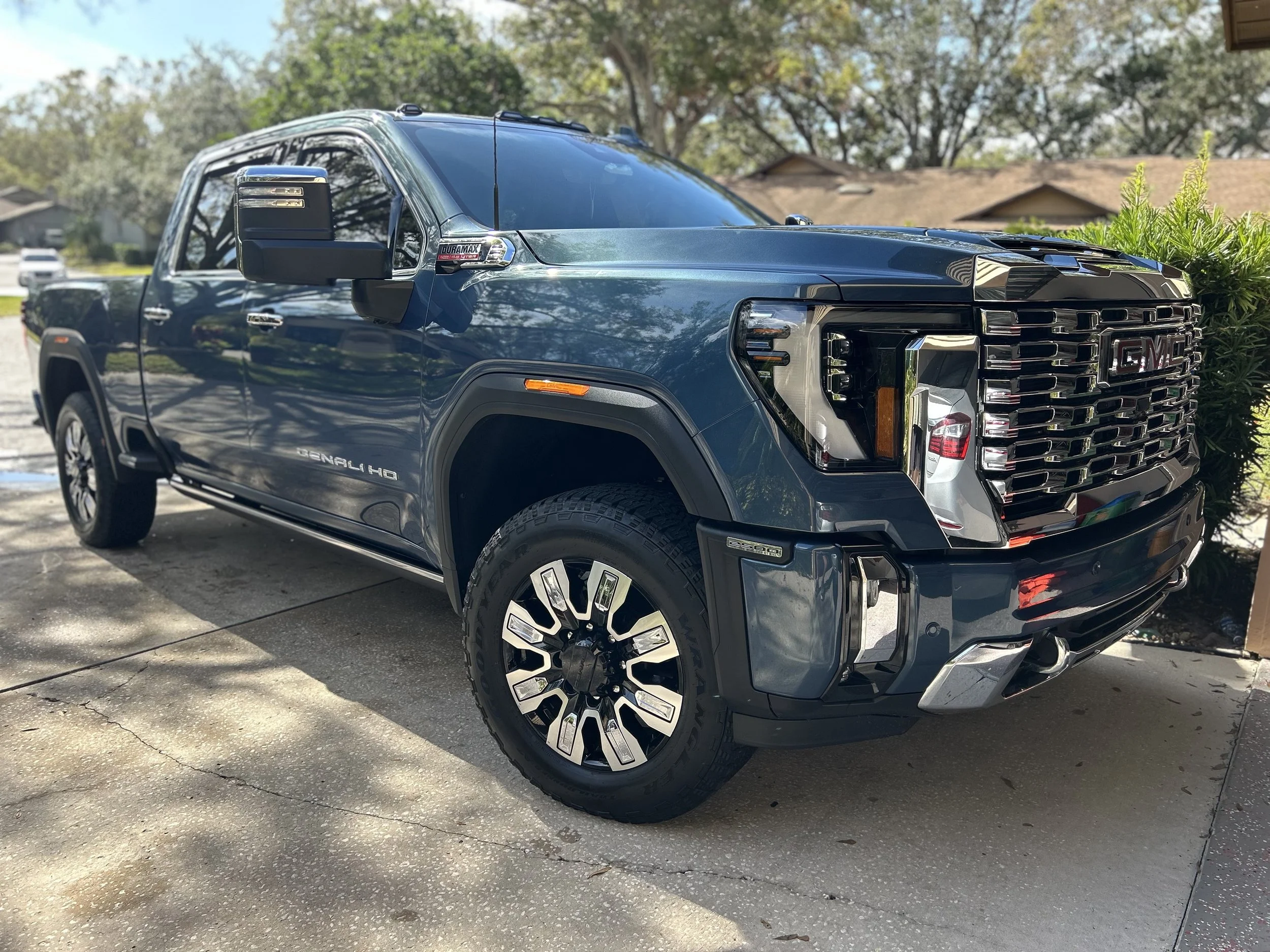 A blue GMC Sierra Denali HD pickup truck parked on a driveway with trees and houses in the background.