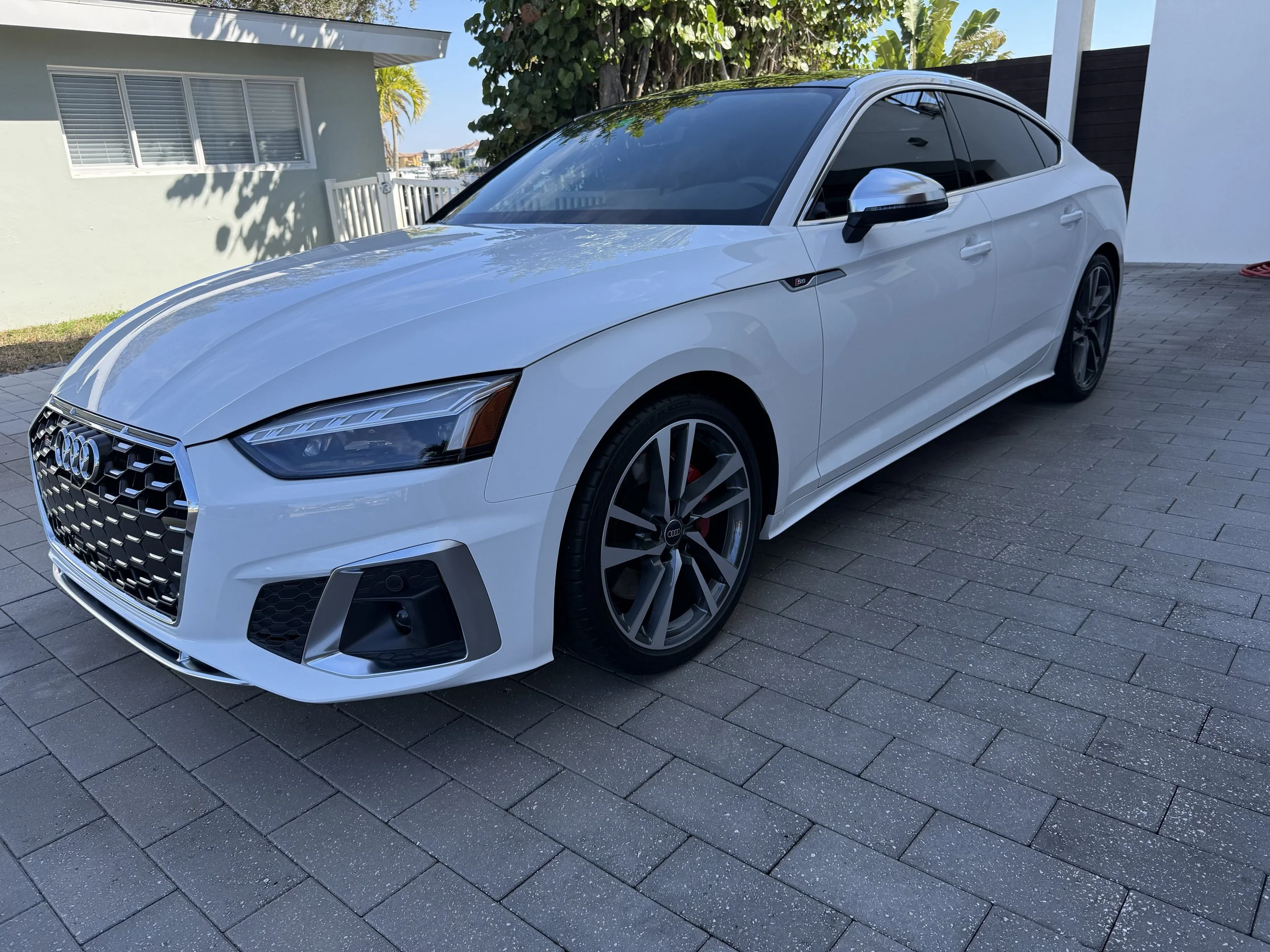 White Audi sedan parked on a paved driveway outside a house with white walls and windows, with trees and a boat in the background.