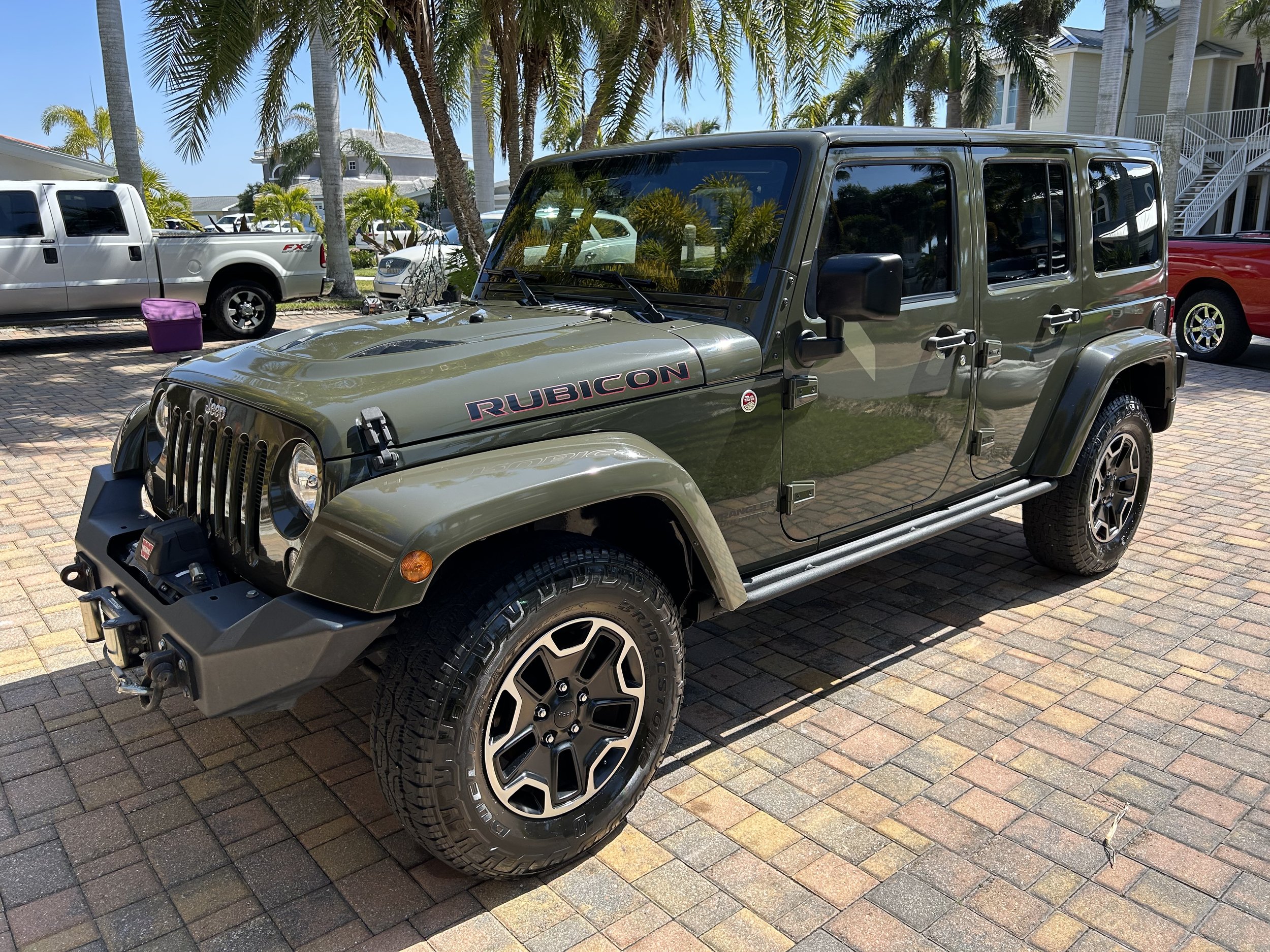 A green Jeep Rubicon four-door SUV parked on a brick driveway, with palm trees and other vehicles in the background.
