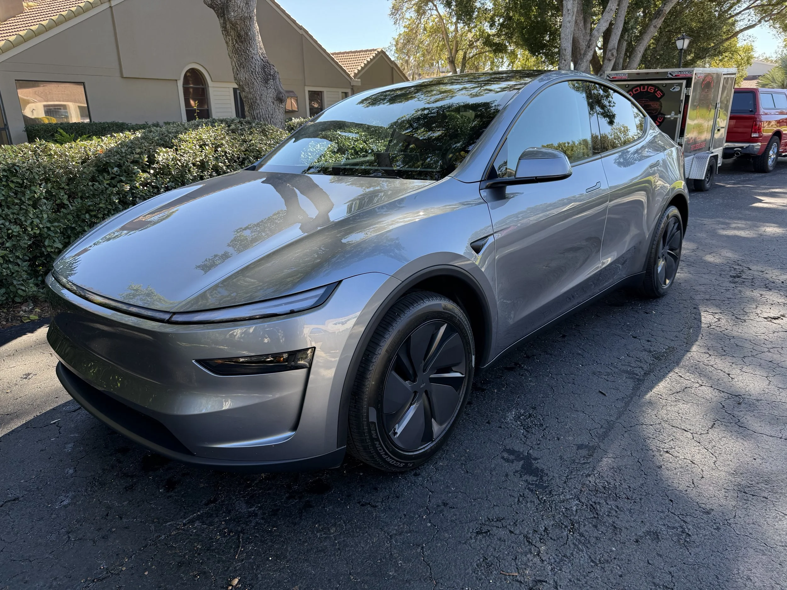 A gray Tesla Model Y parked on a paved street with a hedge and trees in the background, along with a trailer and a red pickup truck.