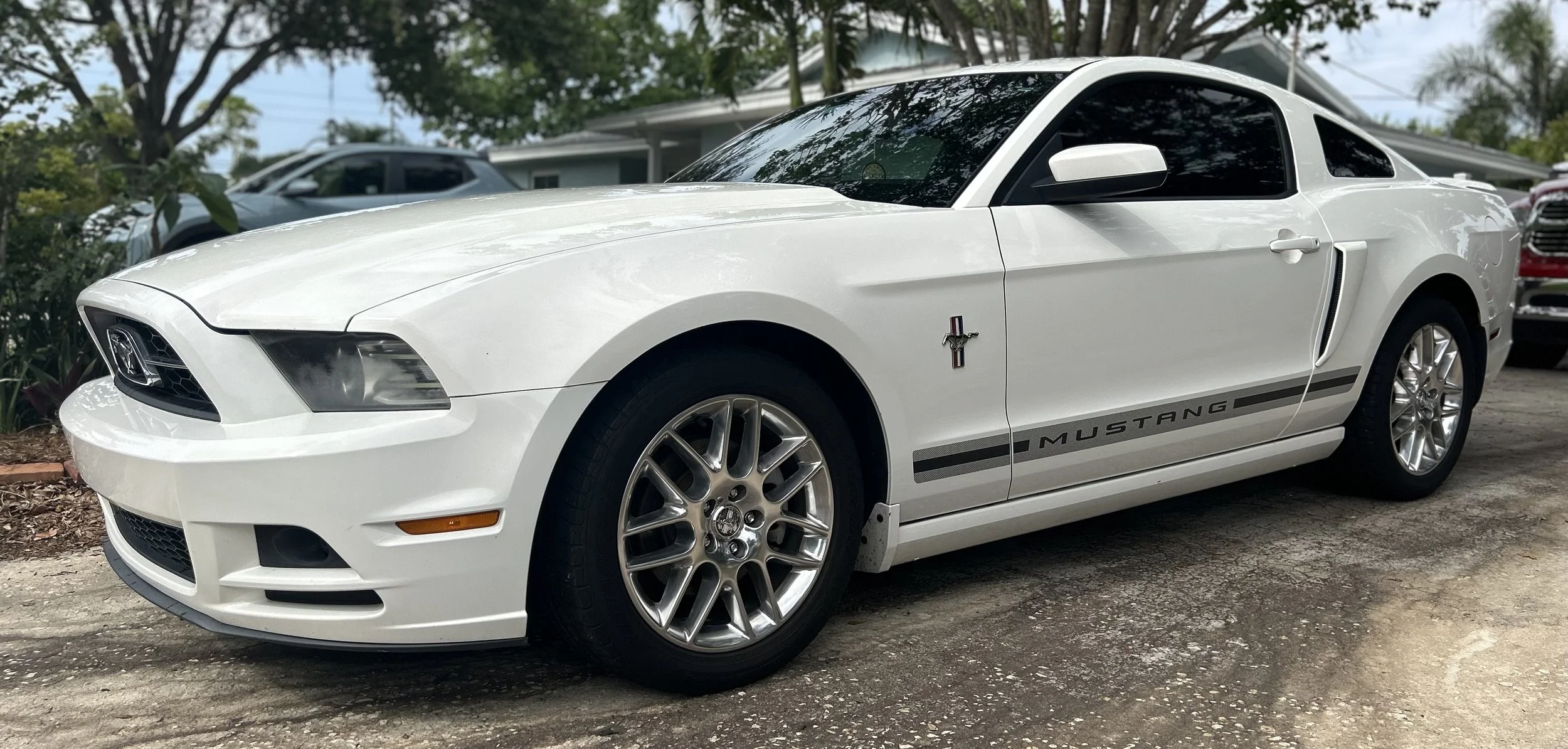White Ford Mustang with black stripe and racing emblem parked on a driveway near trees and other vehicles.