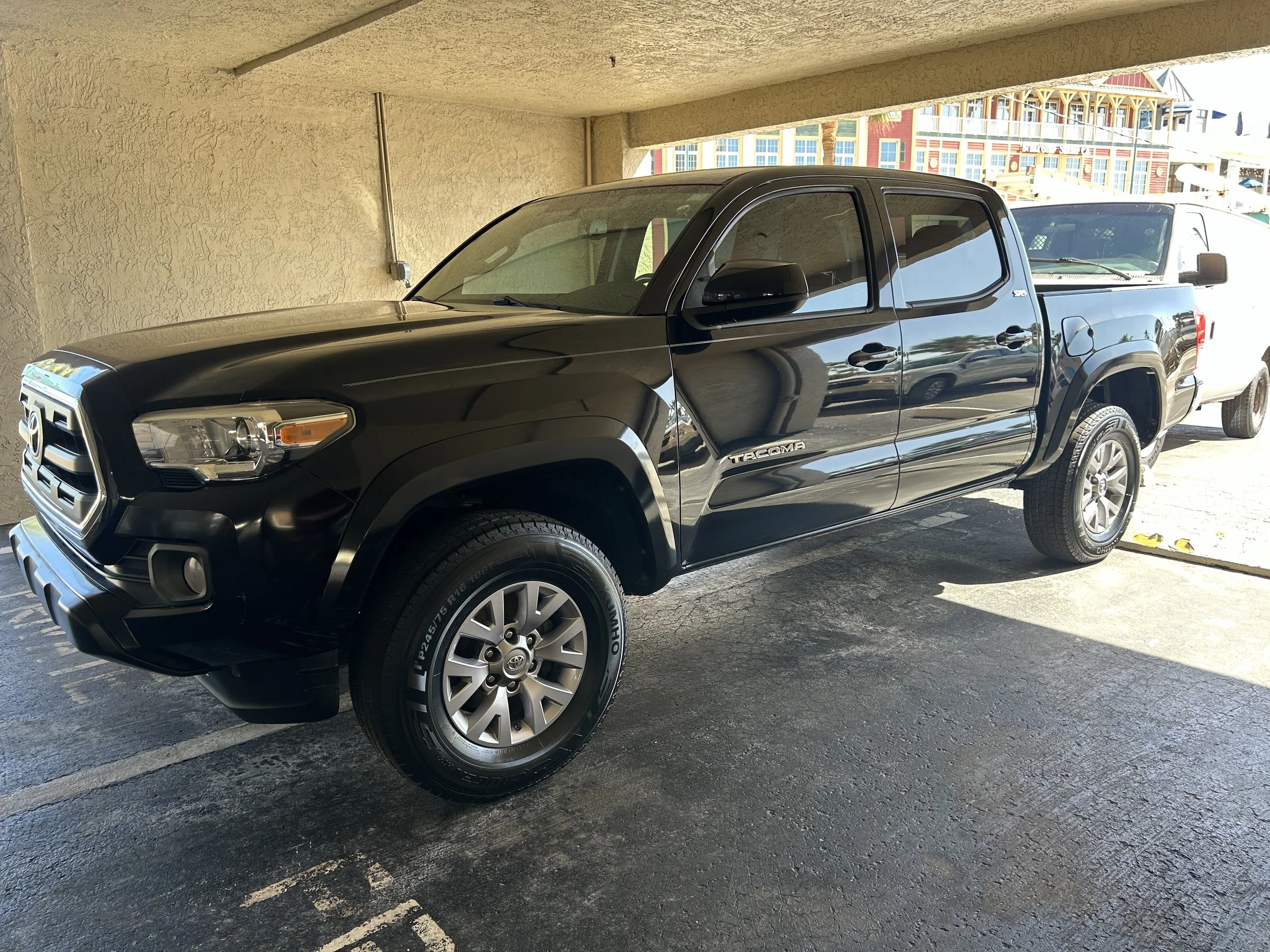 Black Toyota Tacoma pickup truck parked in a covered parking space with a beige wall and a view of colorful buildings in the background.
