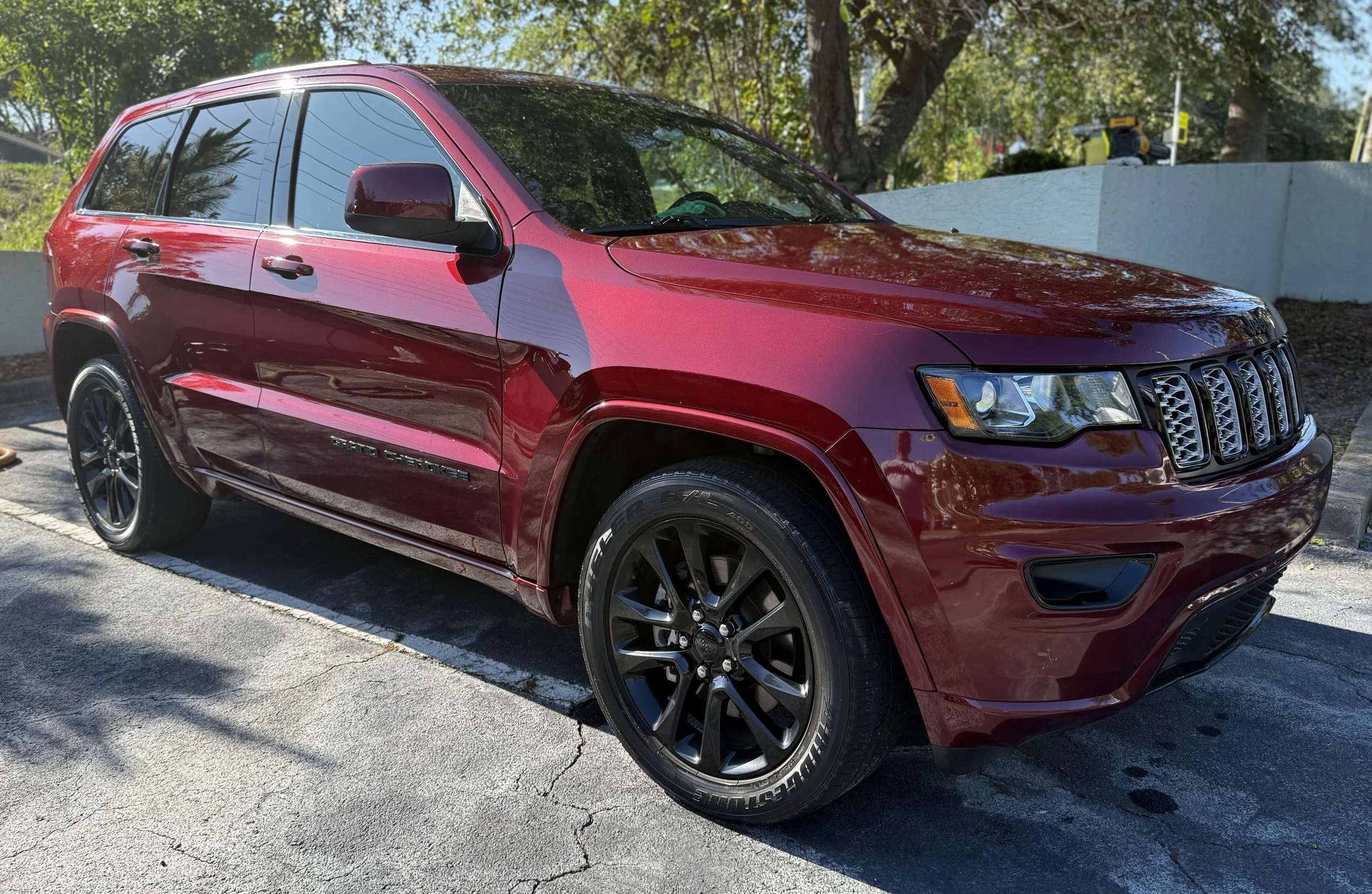 Red Jeep Grand Cherokee parked in a parking lot during daytime, with trees and a white wall in the background.