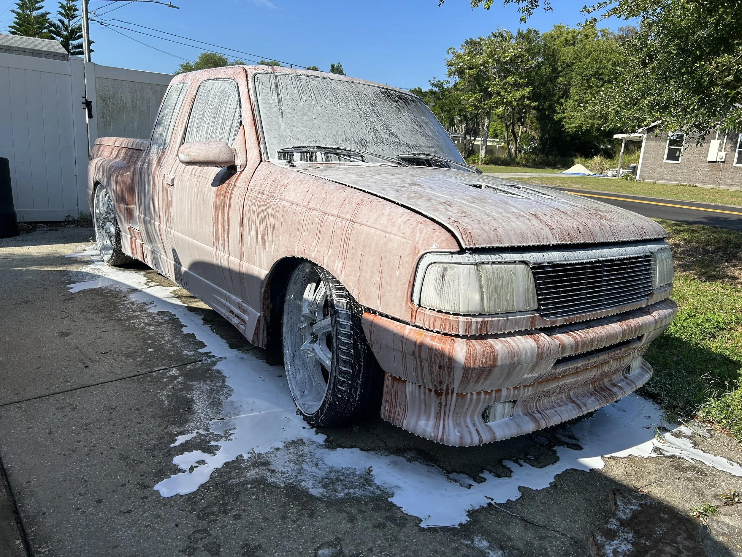 A pink pickup truck covered in soap suds and foam during a wash outside on a sunny day.