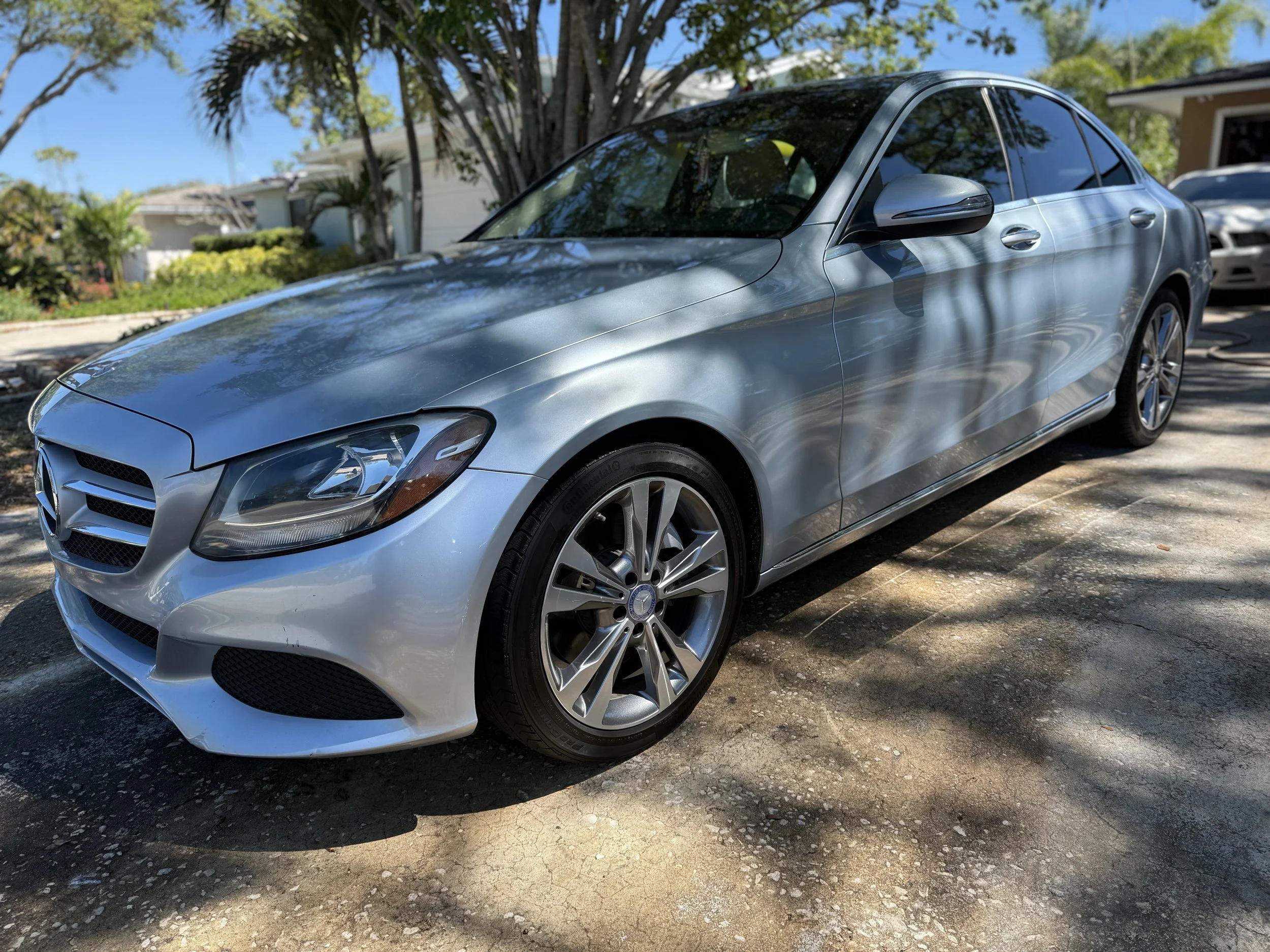 A silver Mercedes-Benz sedan parked on a driveway in front of trees and a house, with reflections of the trees on the car's surface.