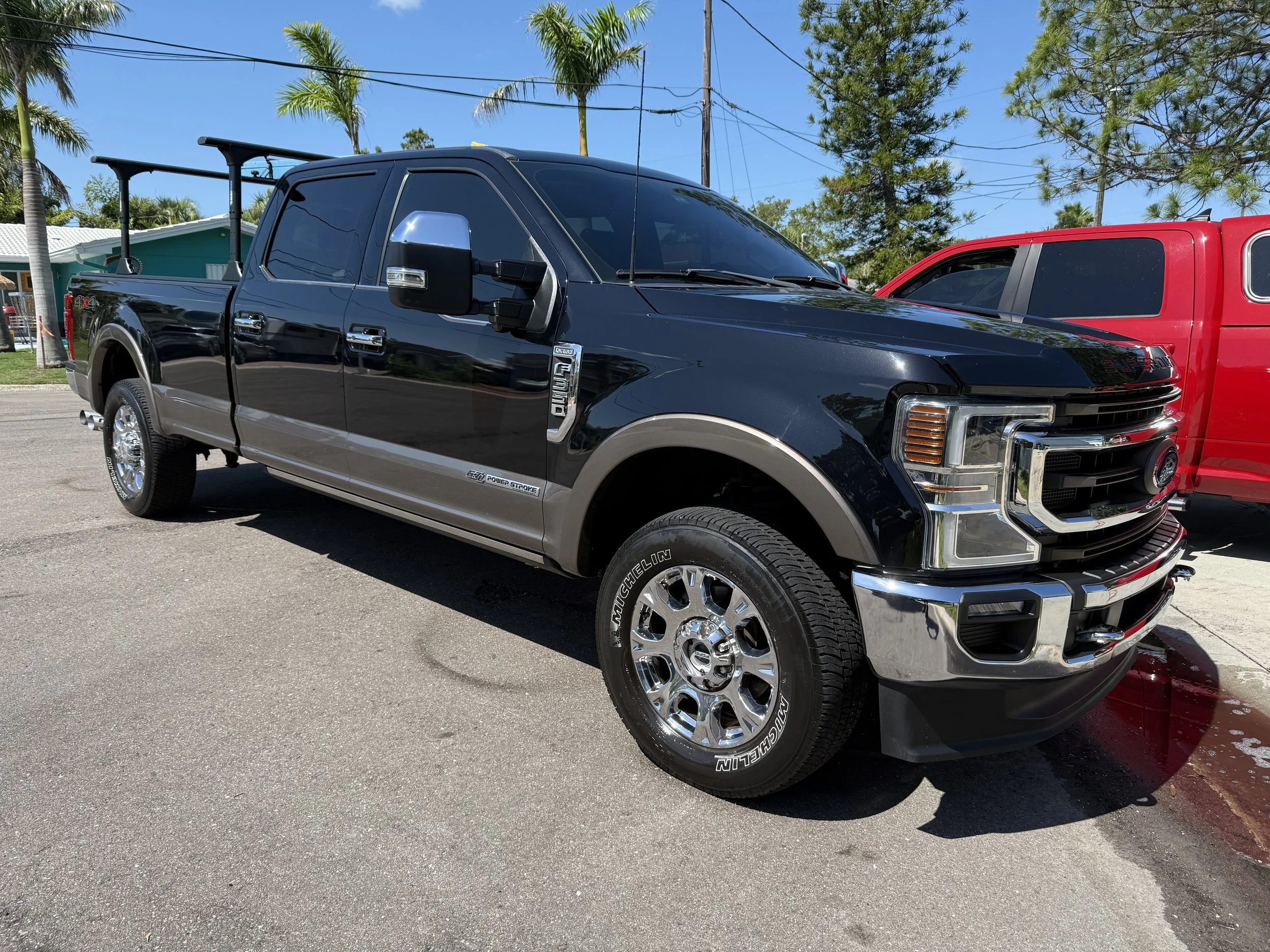 Black and grey Ford pickup truck parked on the street near a red pickup truck, with palm trees and a house in the background.
