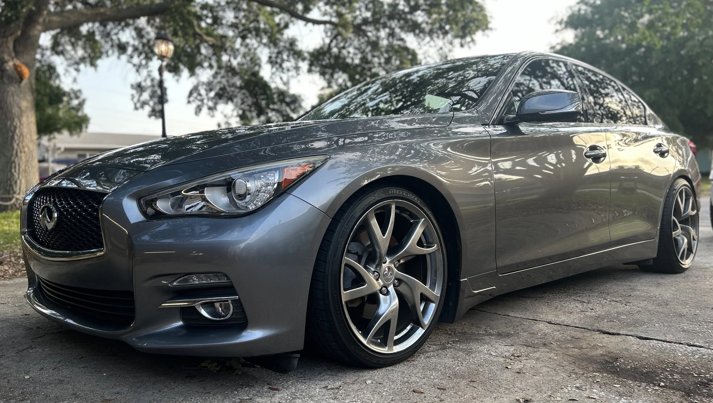 A silver Infiniti G37 coupe parked outdoors on a paved driveway, with trees and a streetlamp in the background.
