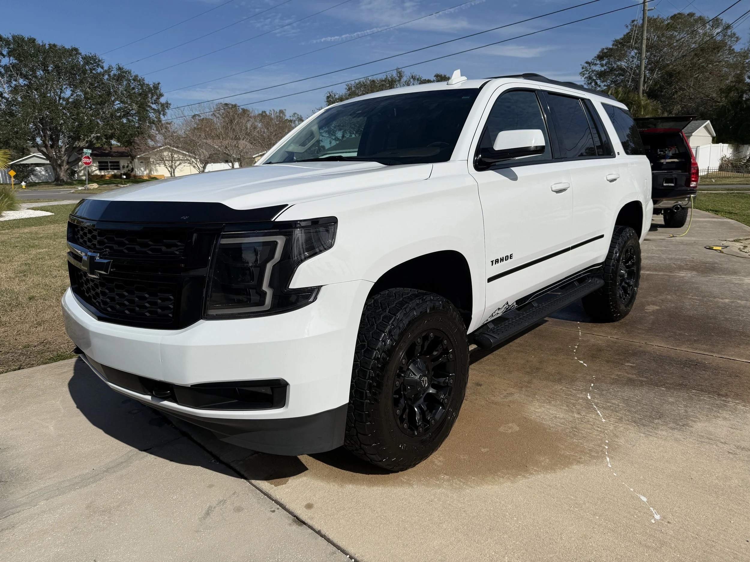 White Chevrolet Tahoe SUV parked on a concrete driveway with a black truck in the background.