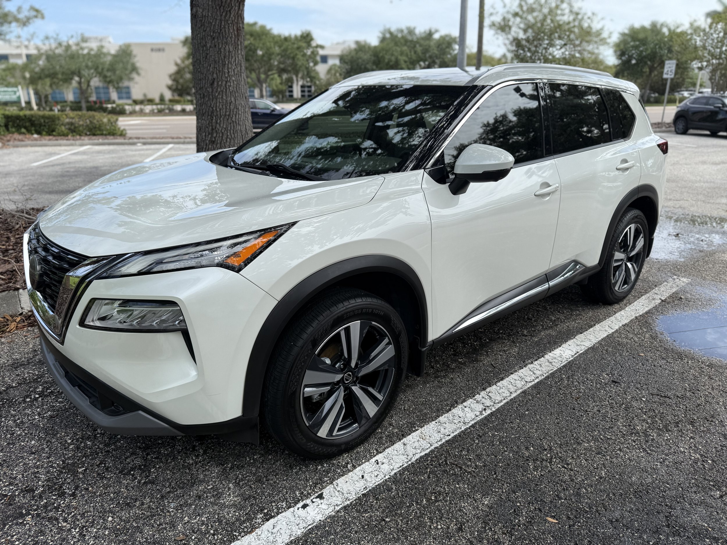 White compact SUV parked in a marked parking spot, with trees and a building in the background.