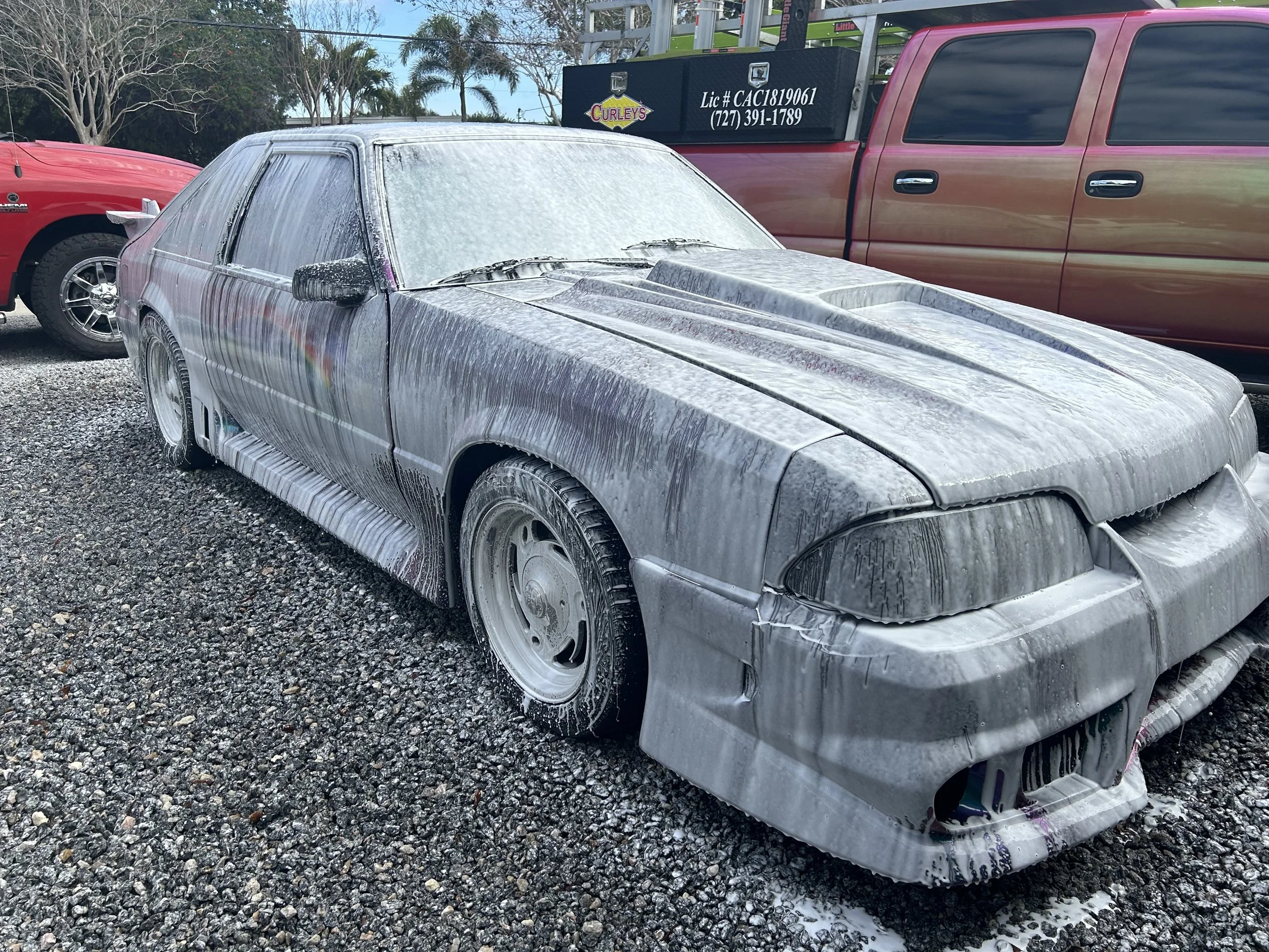 A car covered in white foam, likely from a wash or cleaning process, parked on gravel. The car appears to be a vintage model with a sleek design, surrounded by other vehicles and with palm trees and a cloudy sky in the background.