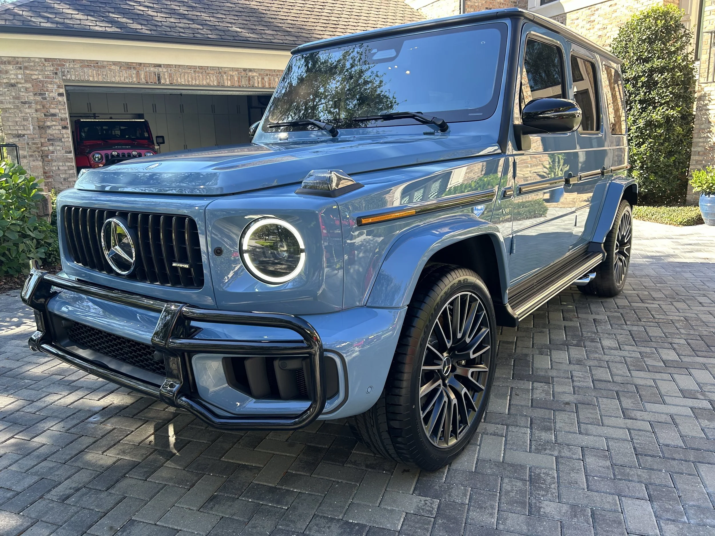 A blue Mercedes-Benz G-Class SUV parked on a brick driveway in front of a house, with trees and plants around.