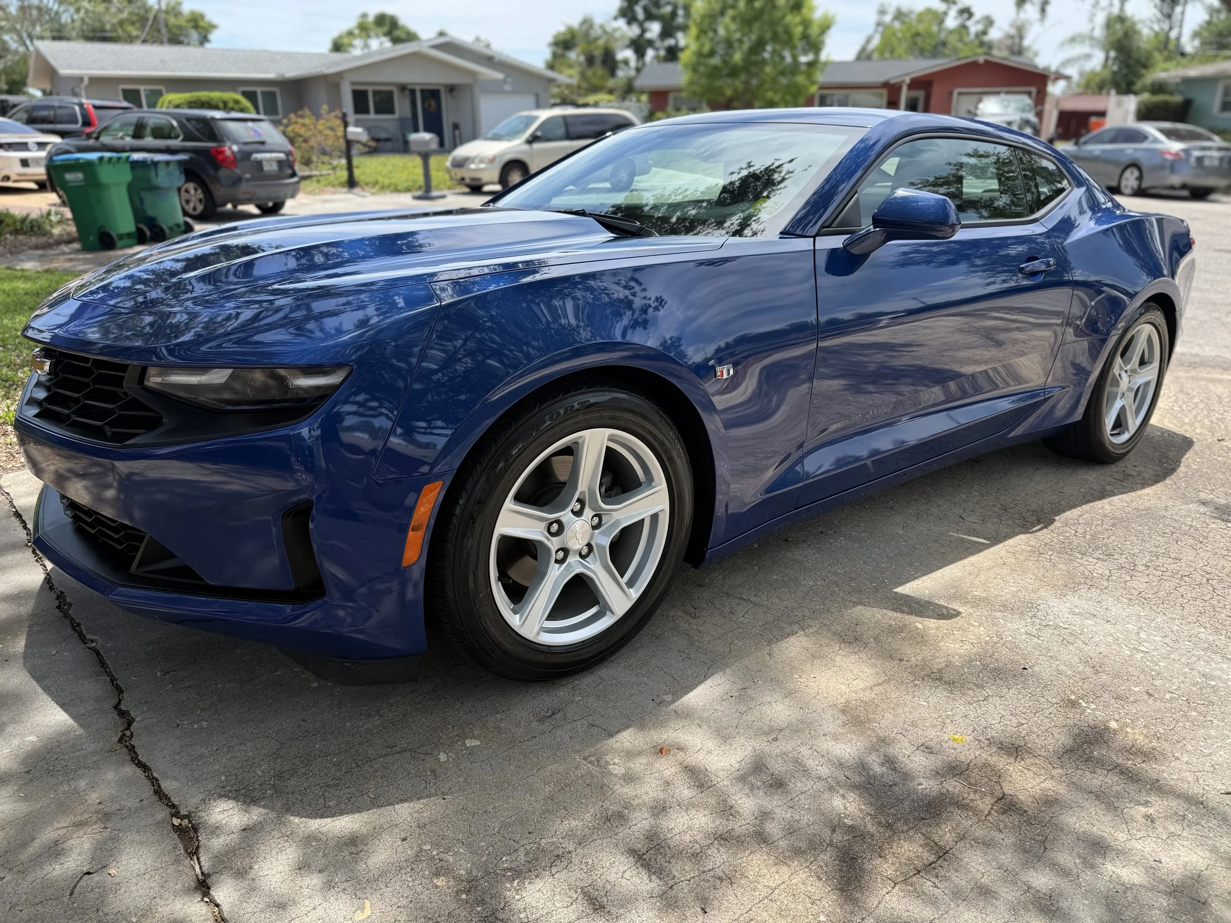 A blue Chevrolet Camaro sports car parked on a residential driveway with houses and trees in the background.