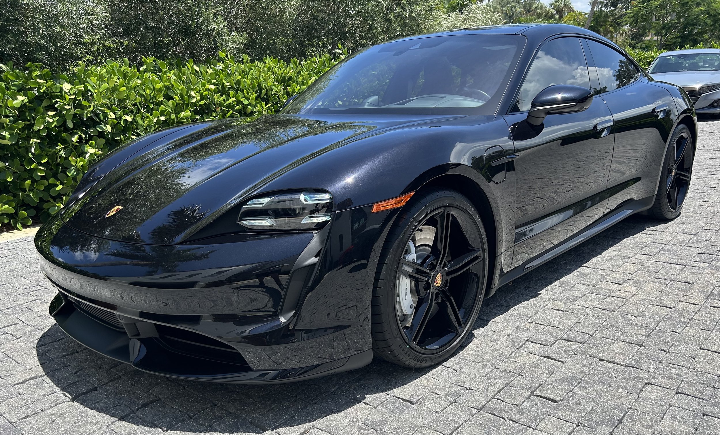 Black Porsche sports car parked on a cobblestone driveway with green bushes and trees in the background.