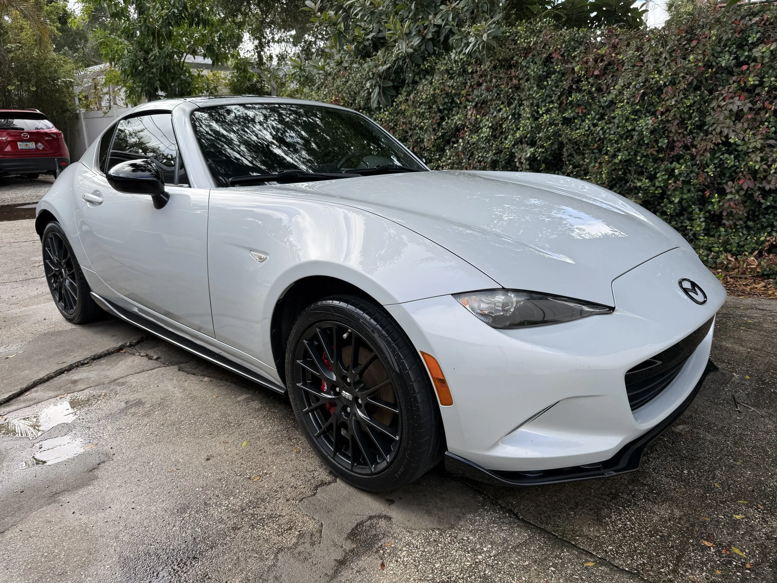 A white Mazda MX-5 Miata convertible sports car parked on a wet driveway with a black front lip spoiler, black wheels, and a black roof. There are trees and bushes in the background.